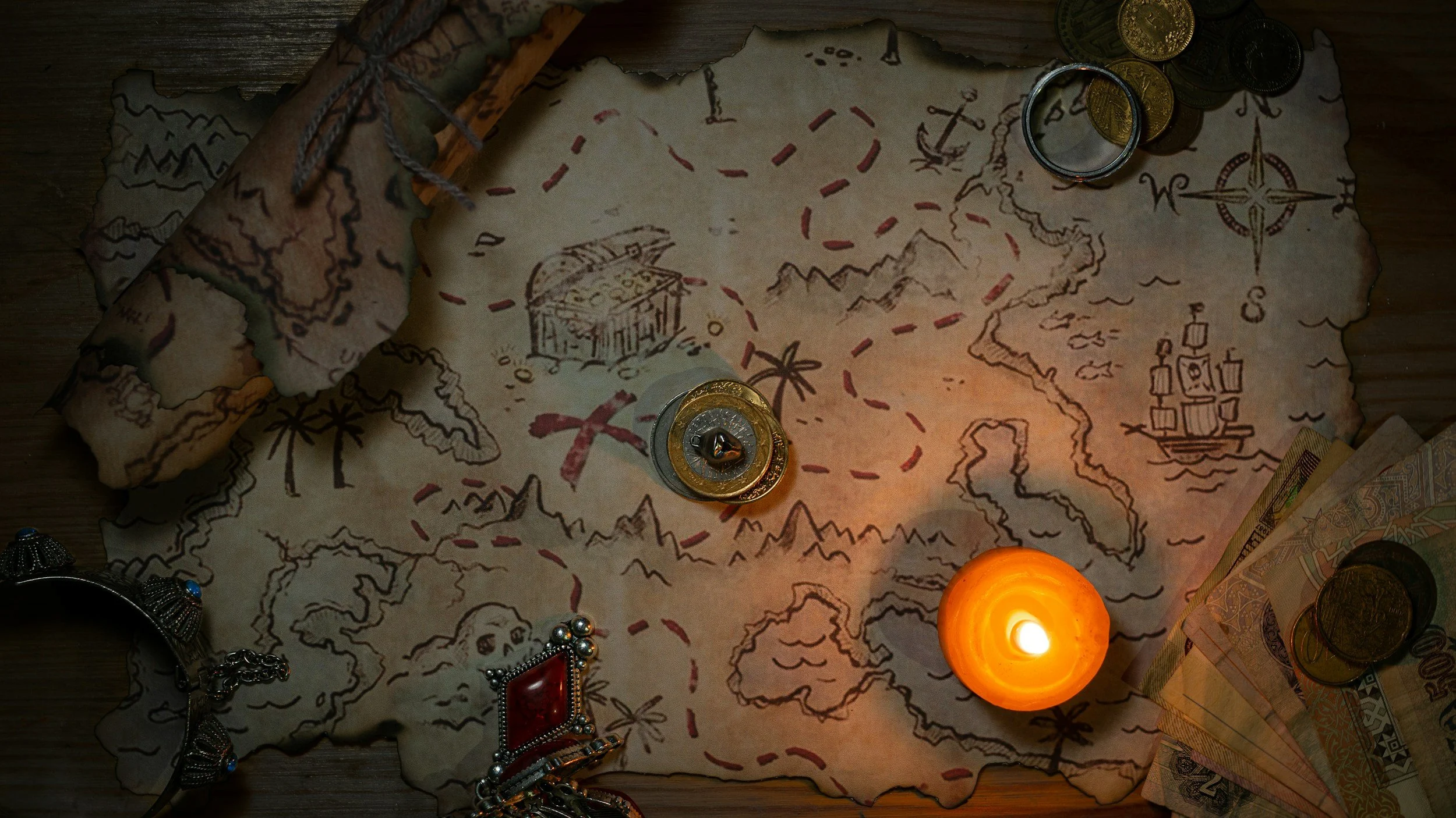 Old treasure map with drawings of an island, a chest, a ship, and a palm tree, surrounded by coins, jewelry, and a lit candle.