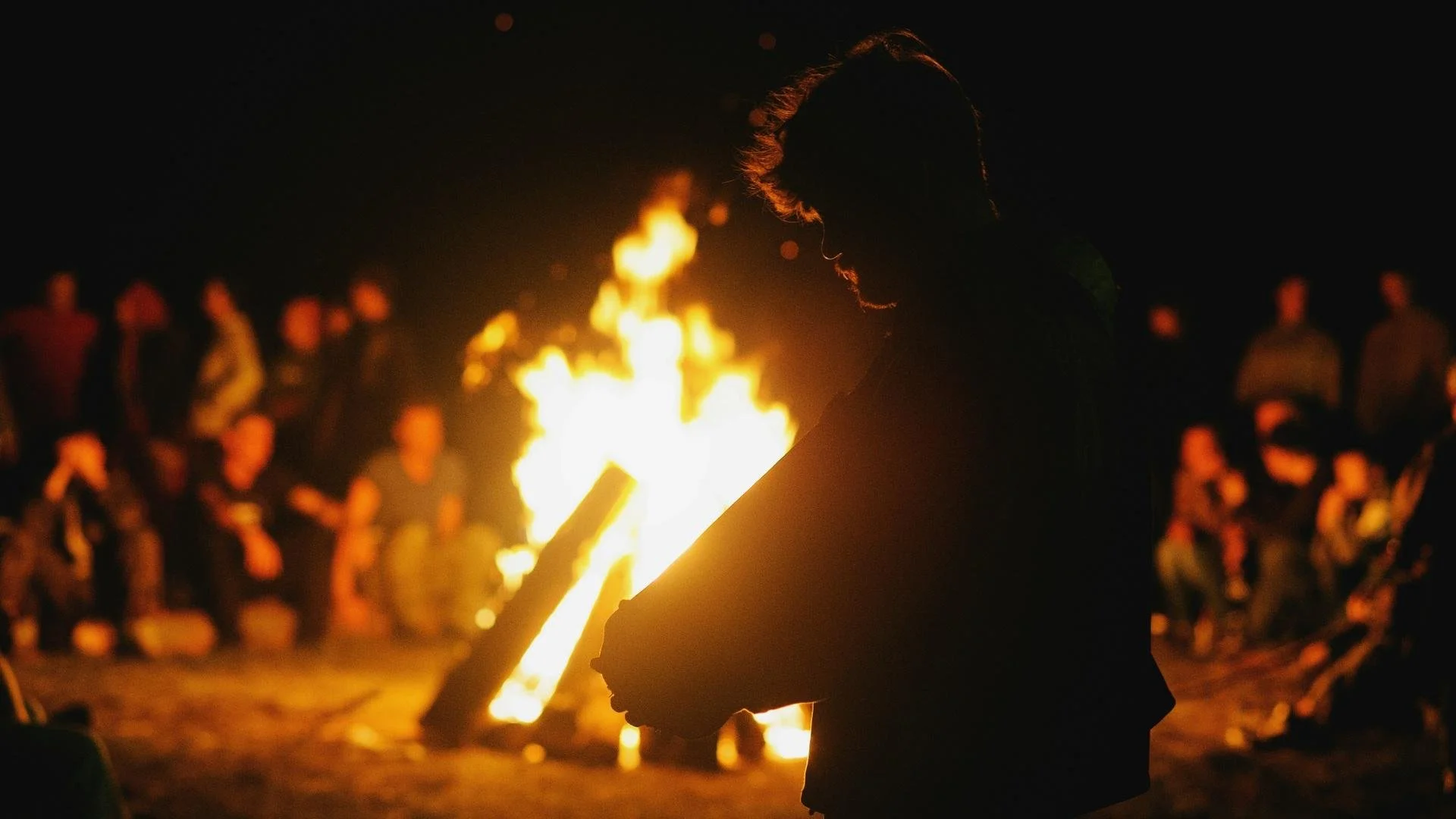 a large bonfire at night in the background illuminating the profile of a man in the foreground