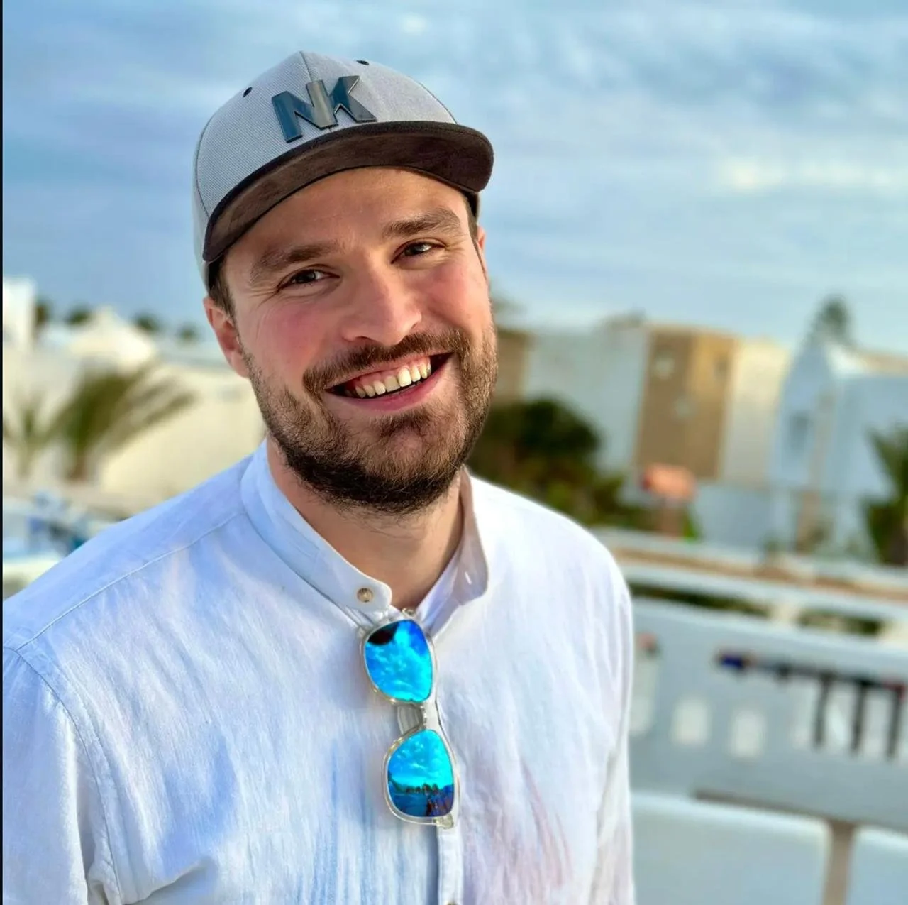 A smiling man with a beard wearing a gray baseball cap with 'NY' on it, sunglasses hanging from his shirt, and a white button-up shirt, outdoors with buildings and palm trees in the background.