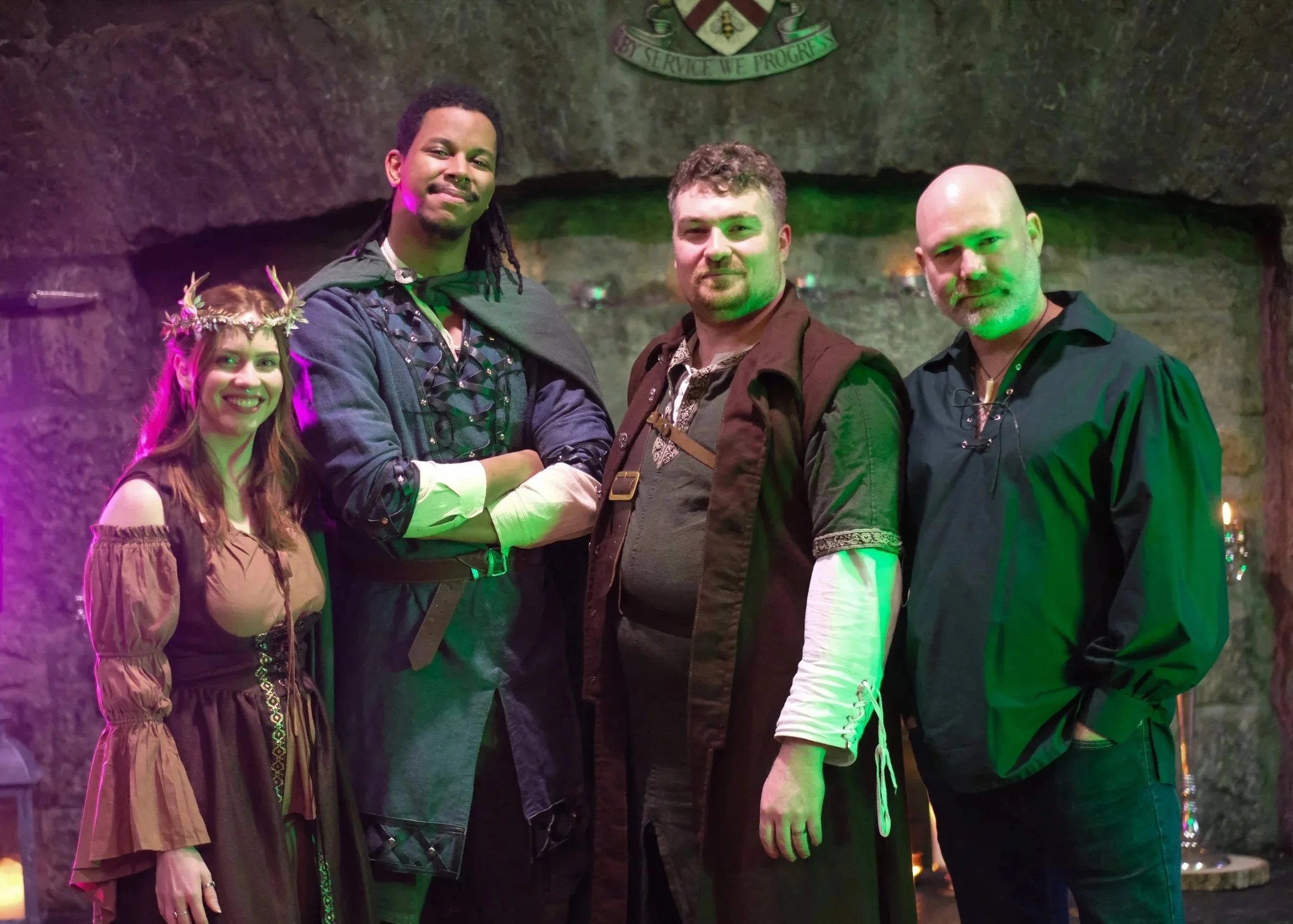 Group of four people standing inside a cave or rock formation. The woman on the left is wearing a brown dress with puffed sleeves and a floral crown. The man next to her has dreadlocks and is wearing a dark jacket with a cape. The man in the middle has short curly hair and is wearing a brown vest over a patterned shirt. The man on the right is bald with a beard and mustache, wearing a dark shirt with a necklace. There is a crest and a banner with the motto 'Service We Progress' on the cave wall behind them. Colored lights illuminate the scene.