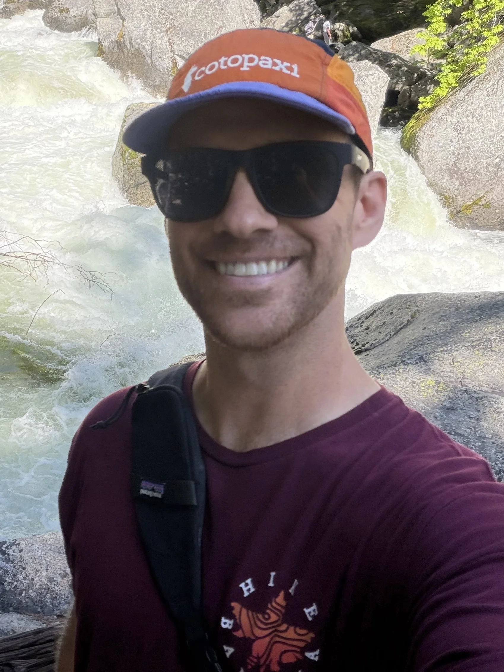 A smiling man wearing sunglasses, a baseball cap, and a maroon shirt, standing in front of a rushing river with rocks and green foliage.
