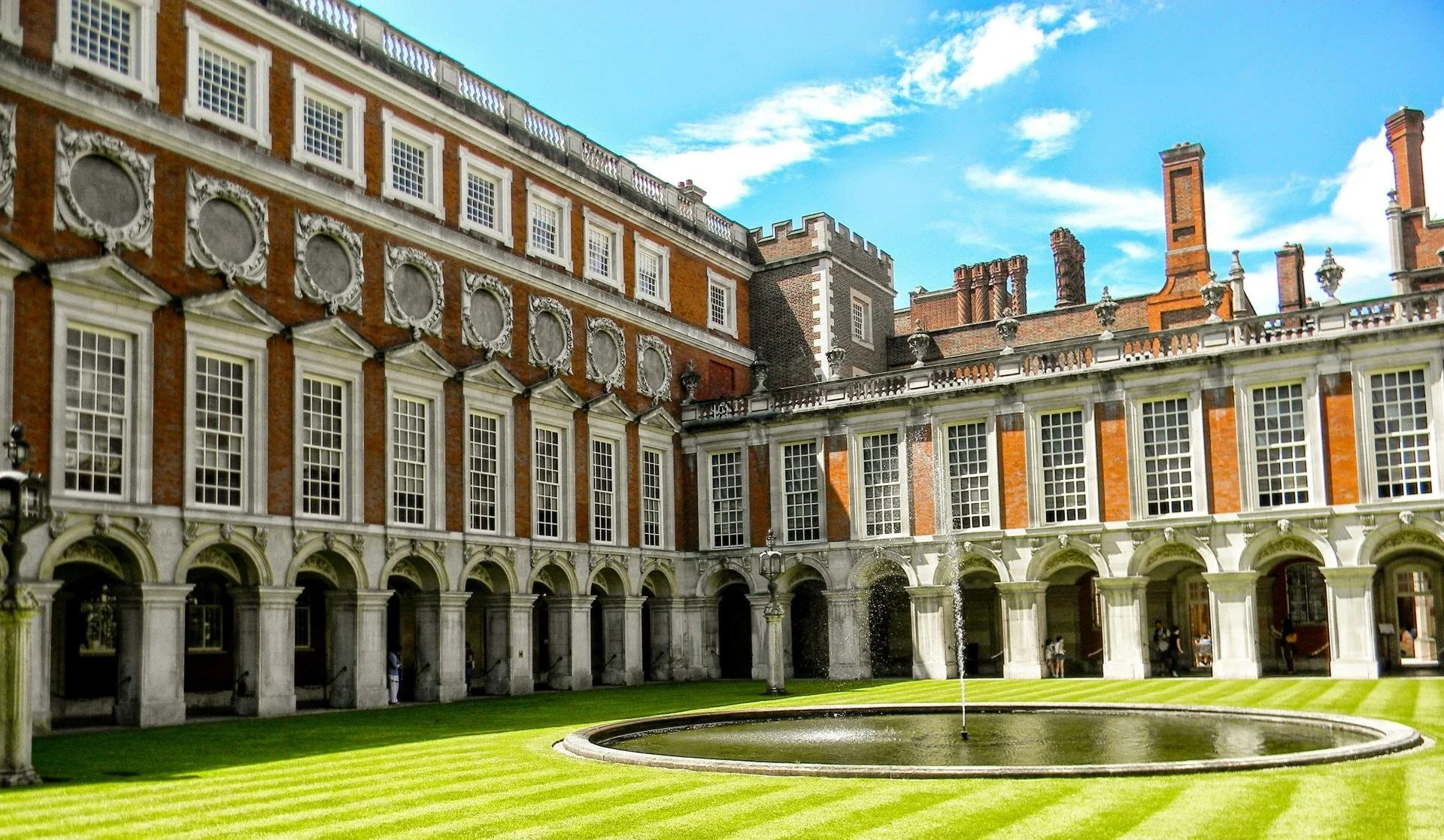 the interior of a courtyard in hampton court palace; rich green grass and bright red brick  around many white arches and window fittings