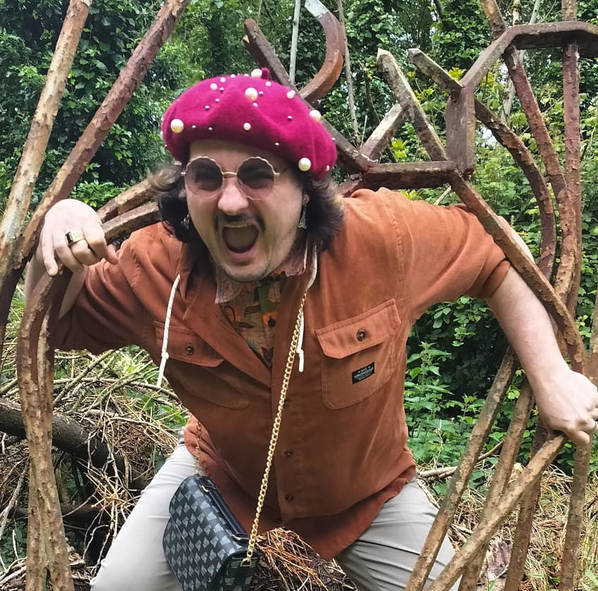A man in sunglasses and a maroon beret with white pompoms, dressed in an orange shirt, is excitedly posing outdoors among green foliage and twisted rusty metal beams, with a patterned bag hanging from his shoulder.