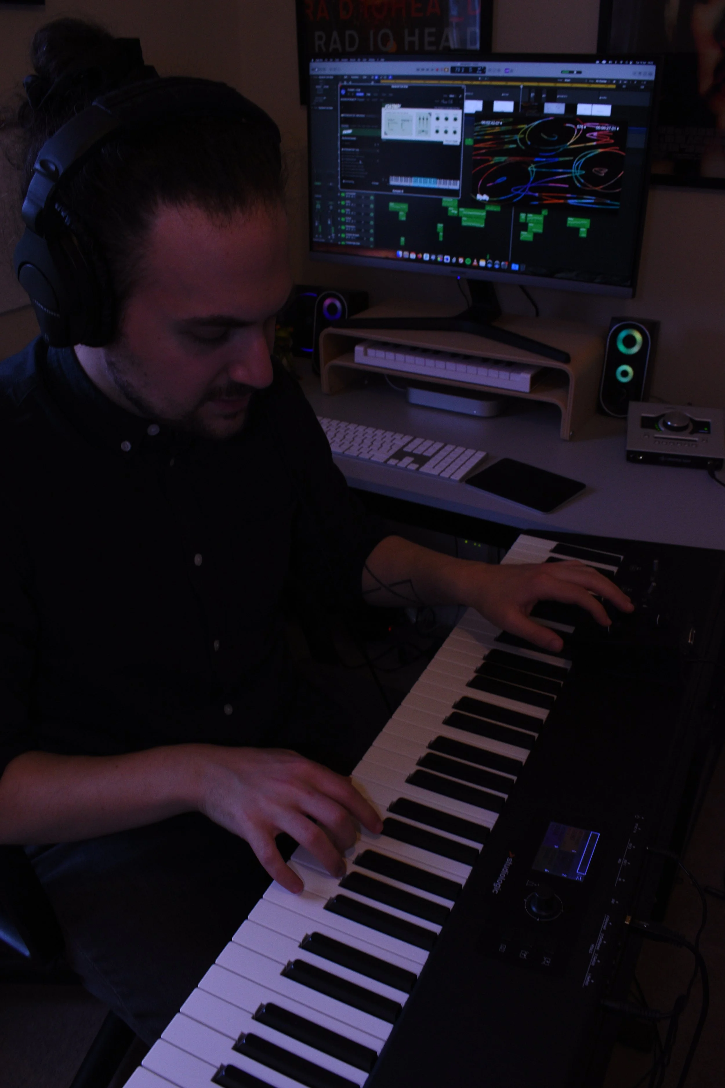 A person wearing headphones playing a keyboard in a dimly lit music studio. A computer monitor in the background displays music production software with colorful waveforms and MIDI notes.