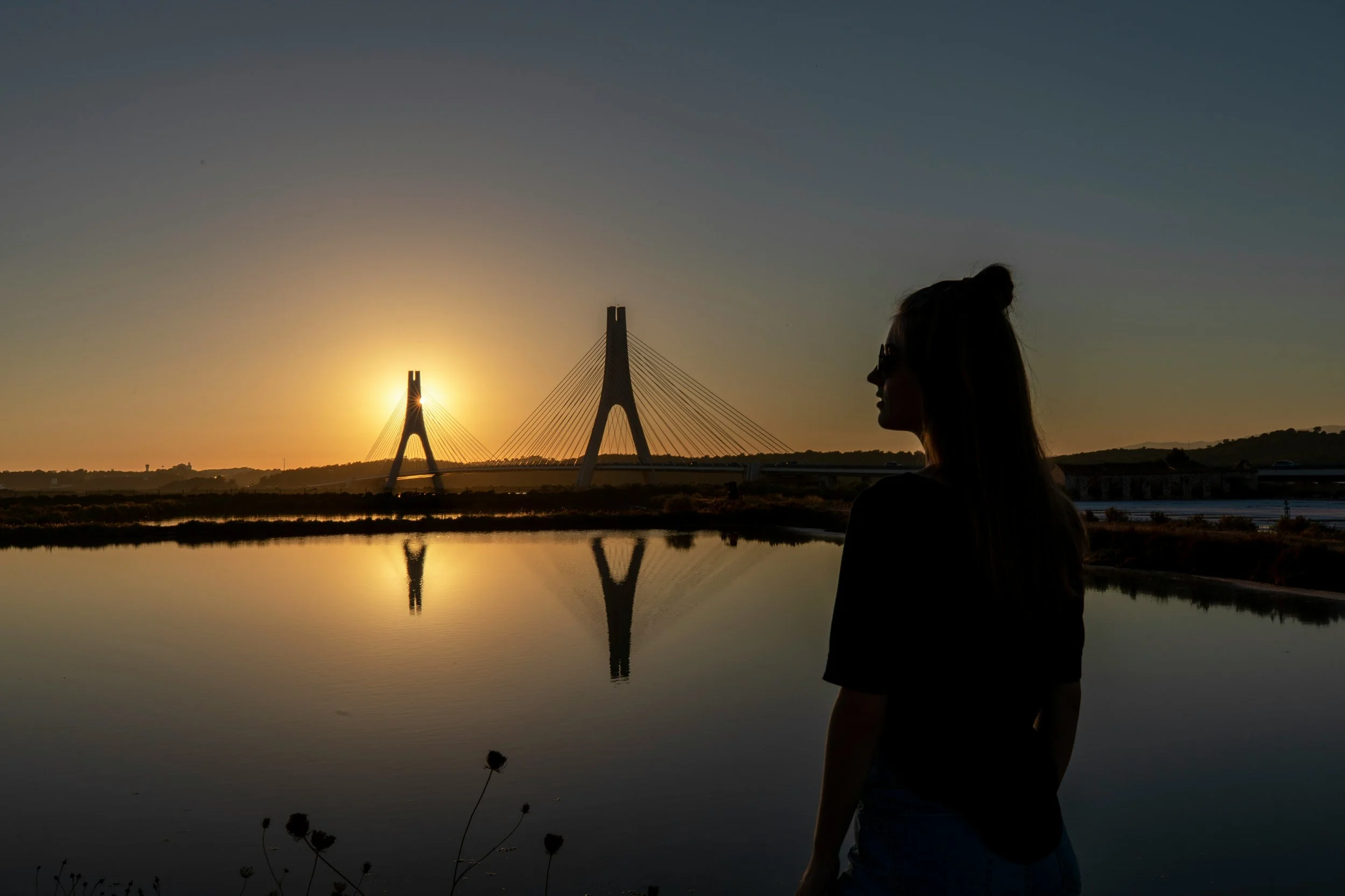 Pessoa de perfil de óculos escuros observando o pôr do sol sobre uma ponte moderna refletida em um rio calmo.