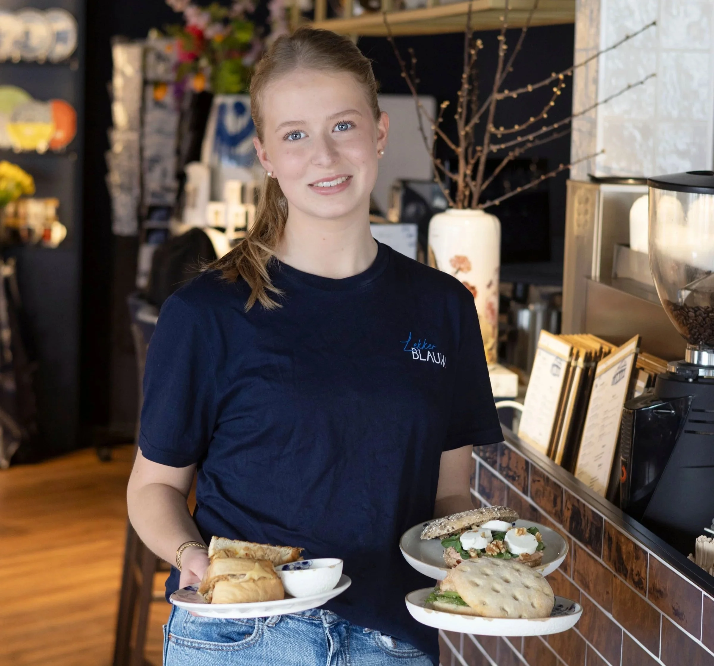 Vrouw met lang haar in eenBlauw t-shirt, die een dienblad met eten vasthoudt, in Lekker Blauw in Putten en Delft. Een eigentijds Lunch concept gefocust op Nederlandse traditie en cultuur.