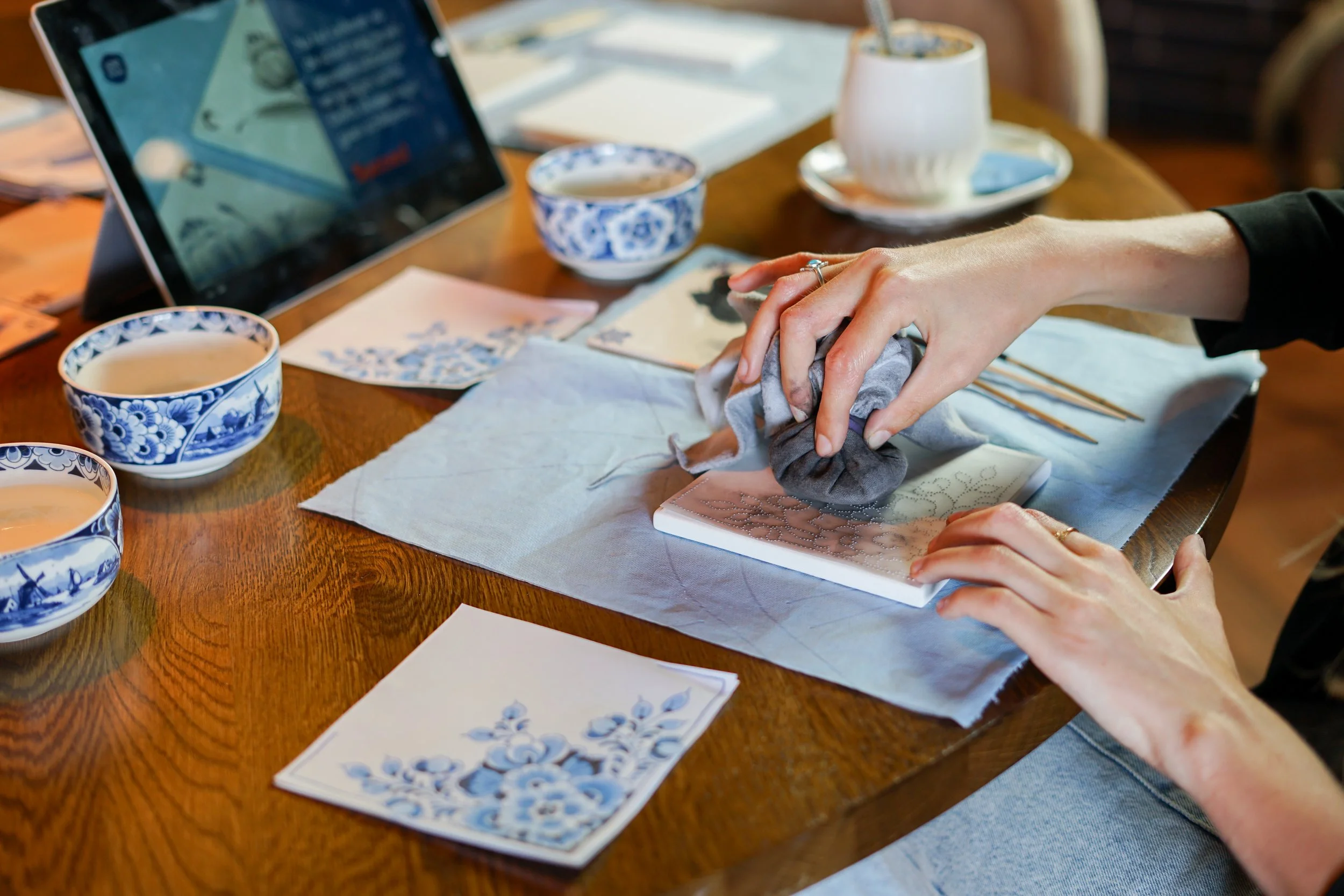 Persoon die keramiek beschildert met blauwe en witte patronen, omringd door kopjes en tekenmaterialen op een houten tafel.