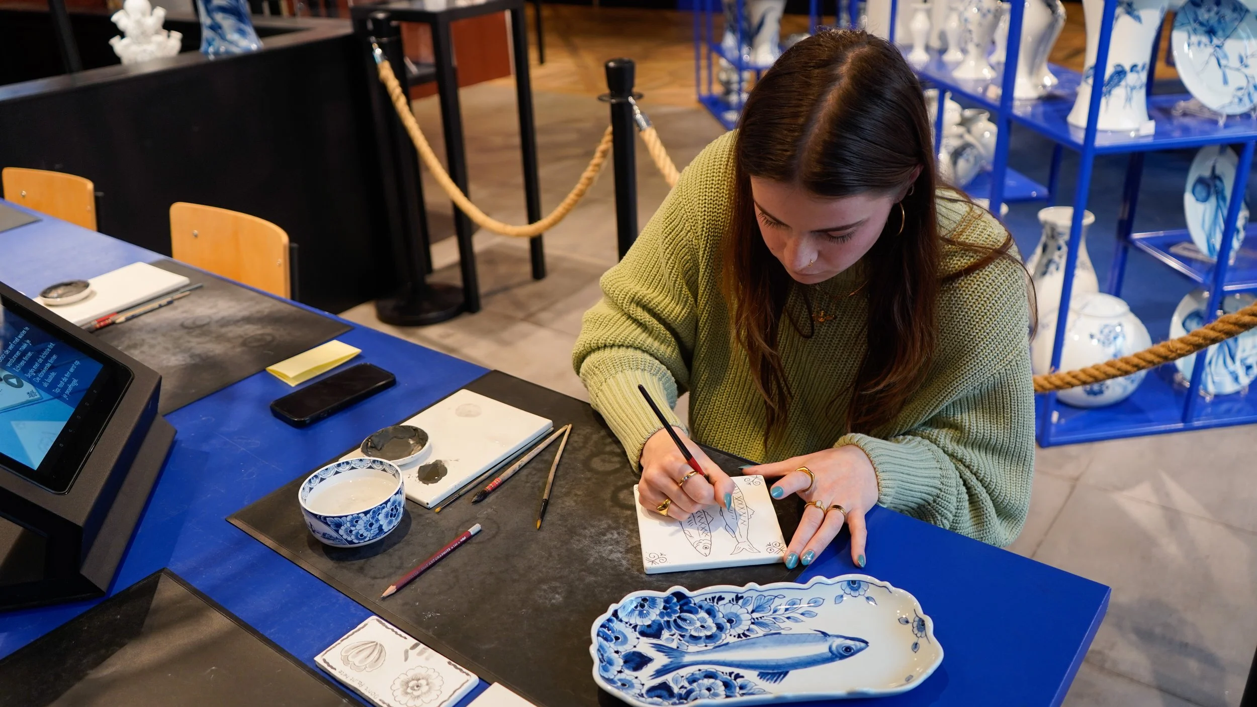Vrouw die bezig is met het schilderen van een delfts blauwe tegel. Tijden het volgen van een workshop, omringd door schildergereedschap en keramiek objecten in een Heinen Delfts Blauw winkel 