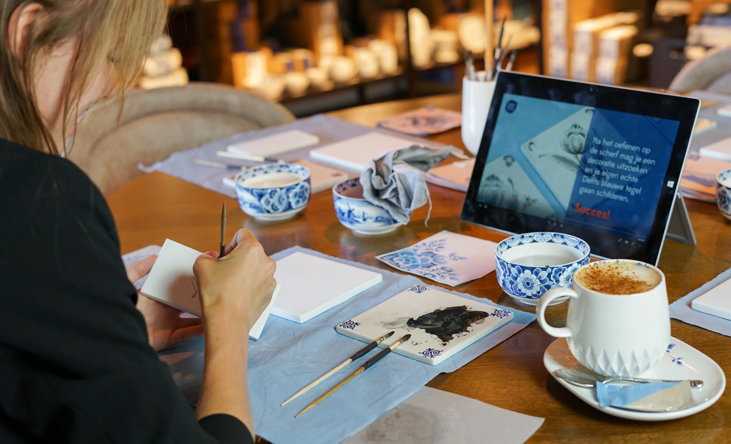 Vrouw die keramiek schildert met blauwe en witte borden en kopjes op een tafel, met een tablet en verschillende schildermaterialen.