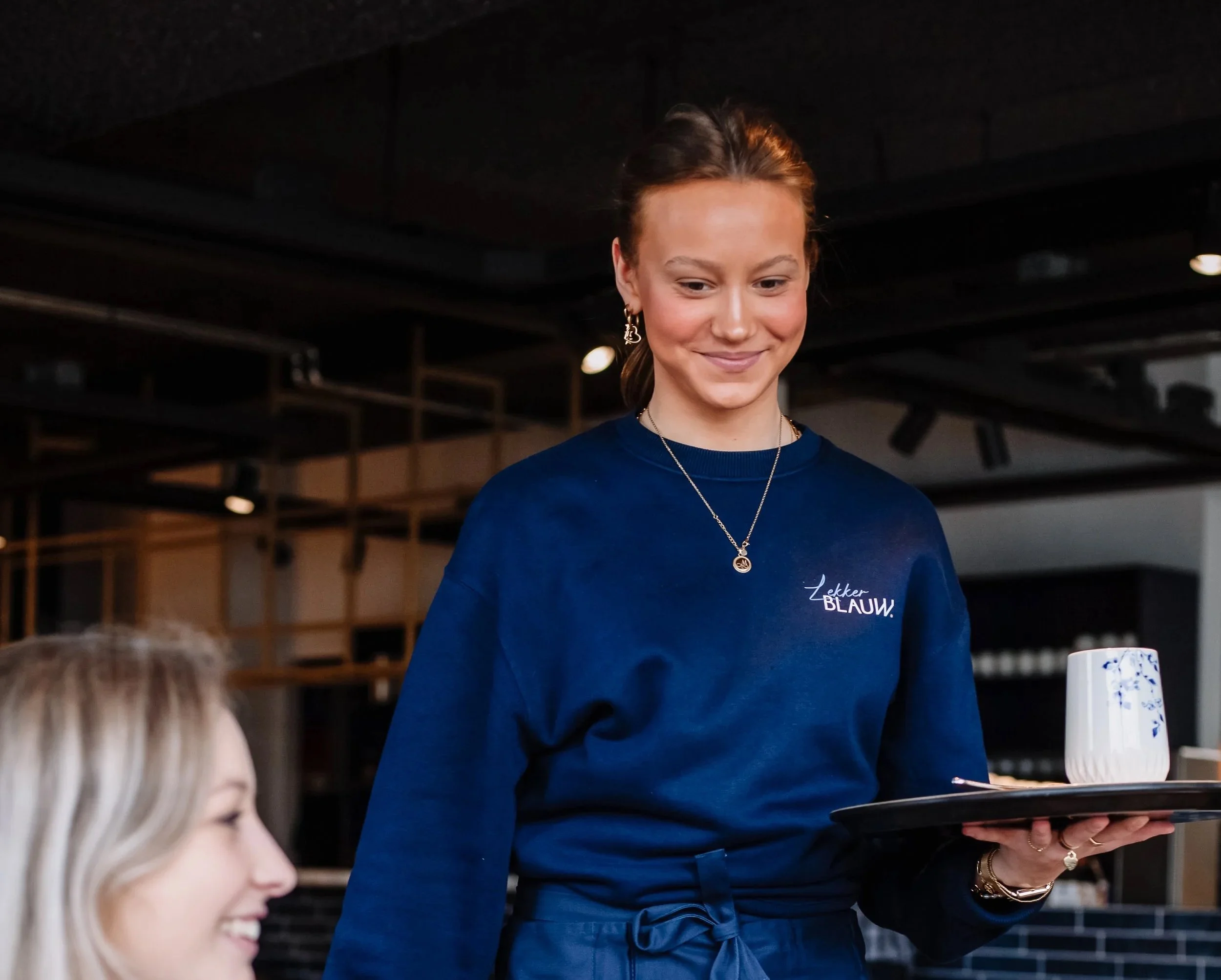 Vrouw met een blauwe trui en parelketting serveert een kopje koffie of thee op een dienblad in een modern restaurant of café.