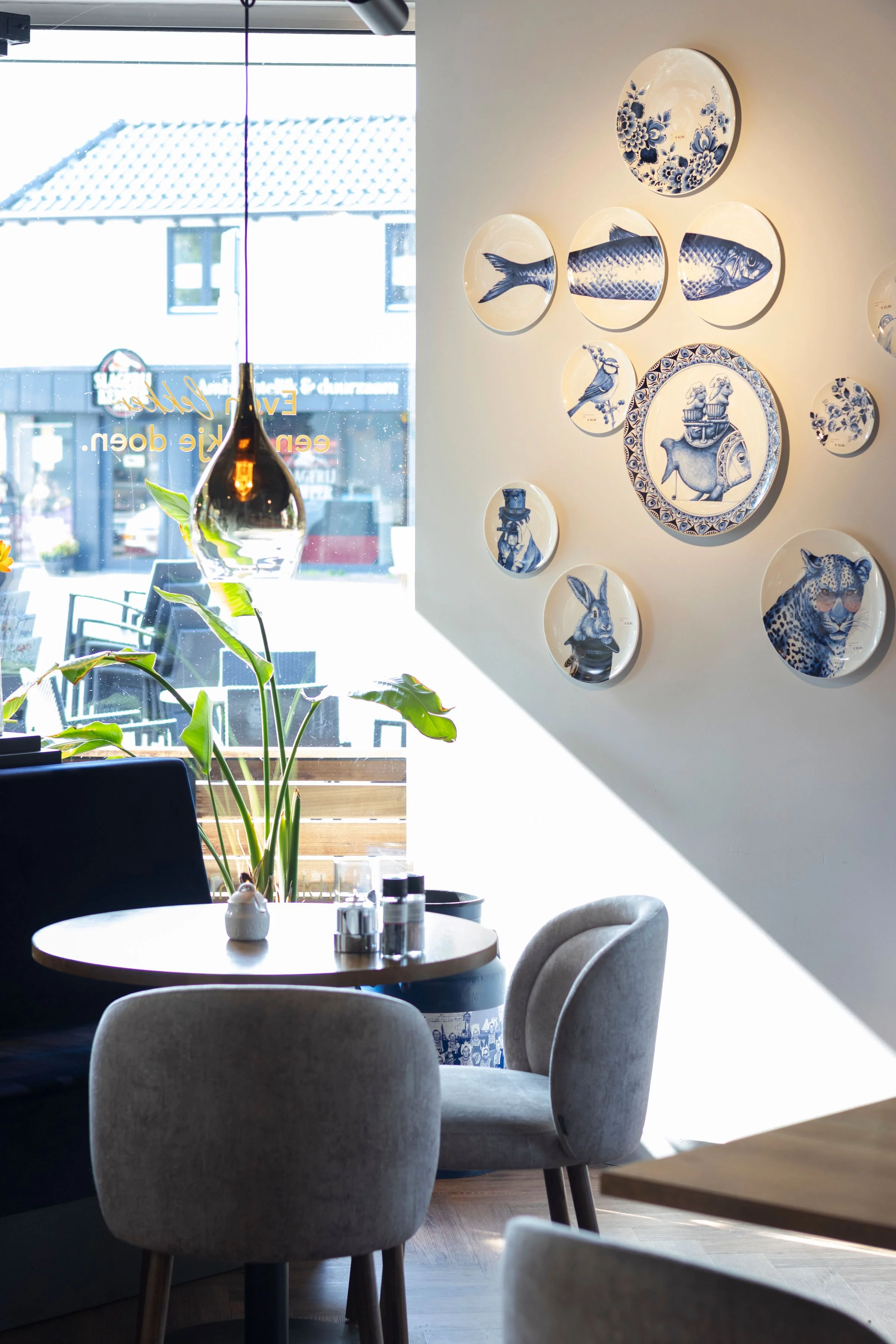Interieur van een café met een ronde tafel, twee stoelen, een plant en muurdecoraties met blauwe patronen van dieren en objecten.