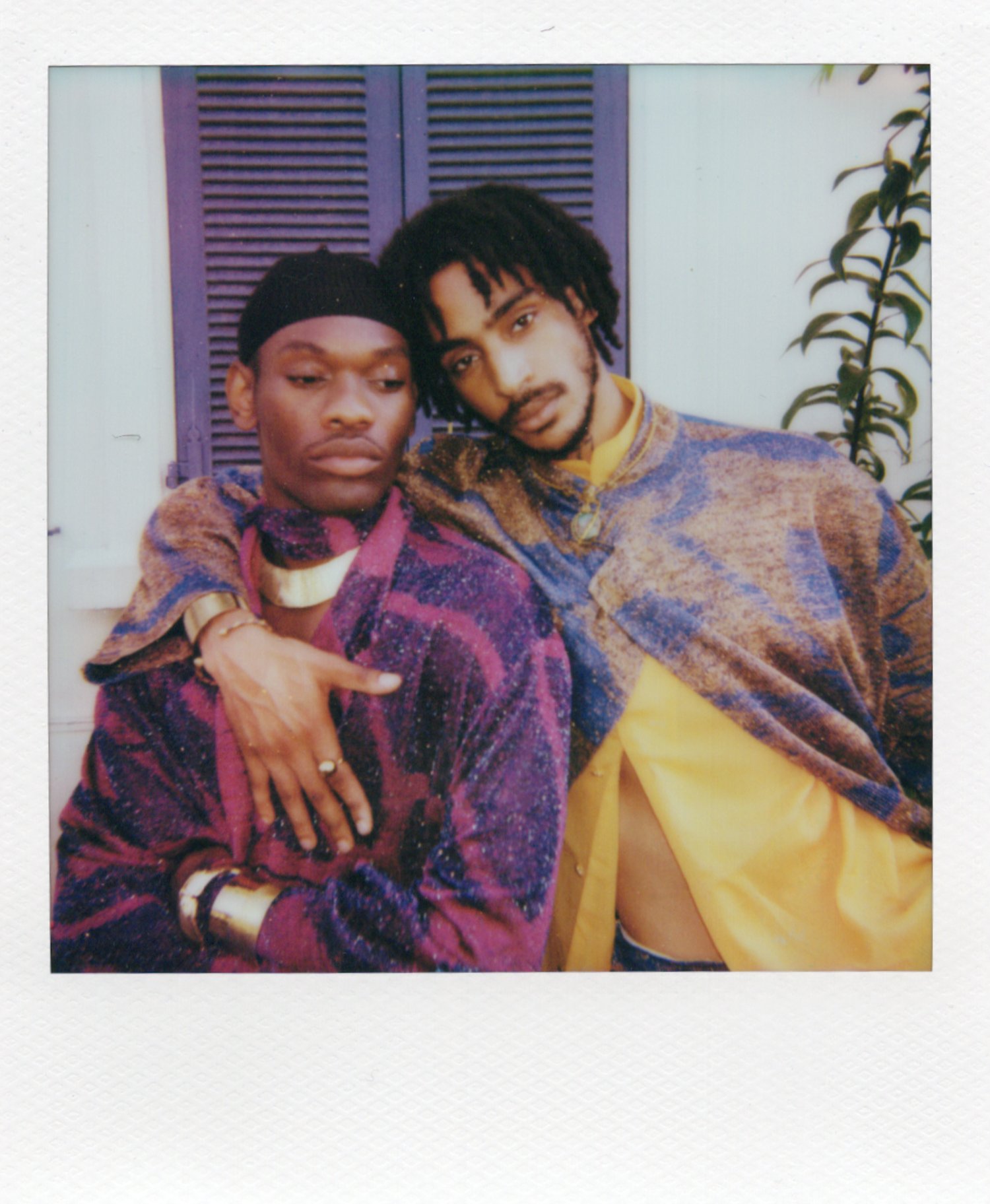 Deux jeunes hommes posant ensemble, vêtus de vêtements colorés avec des motifs, dans un décor avec volets violets et une plante