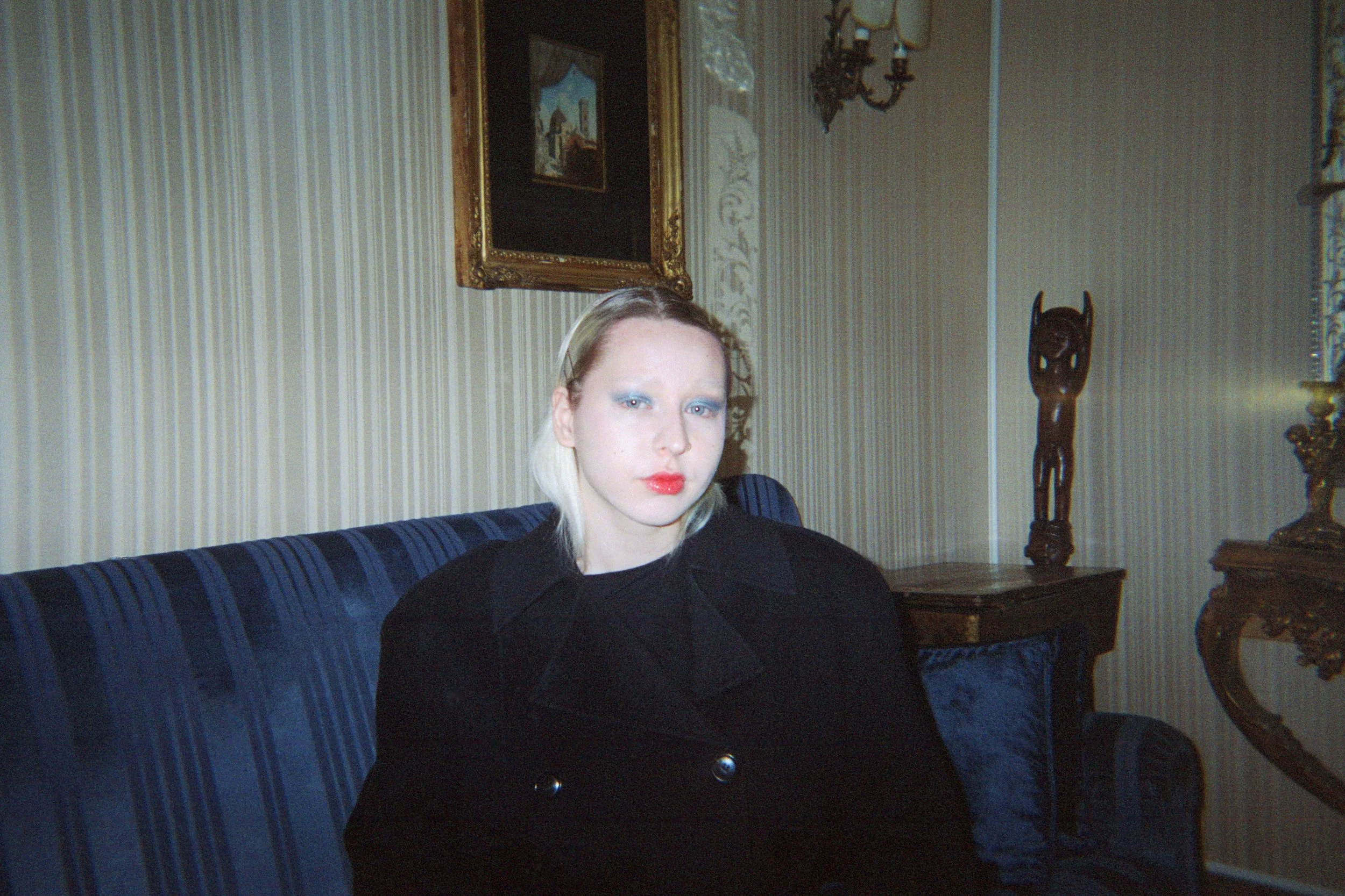 Femme blonde assise sur un canapé en velours bleu dans un salon décoré de manière classique, avec un cadre doré au mur et une sculpture en bois dans le coin.