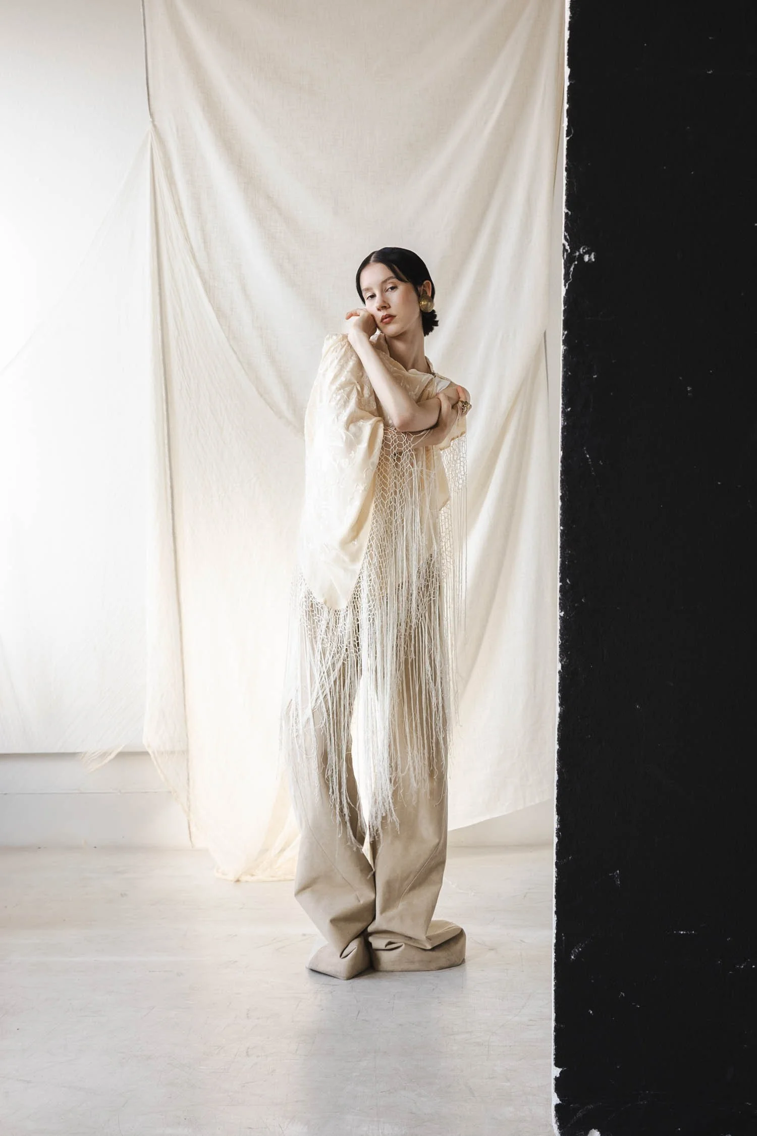 Une femme posant devant un fond beige avec un panneau noir sur le côté droit, portant une tenue élégante de couleur crème avec des boucles d'oreilles en or.
