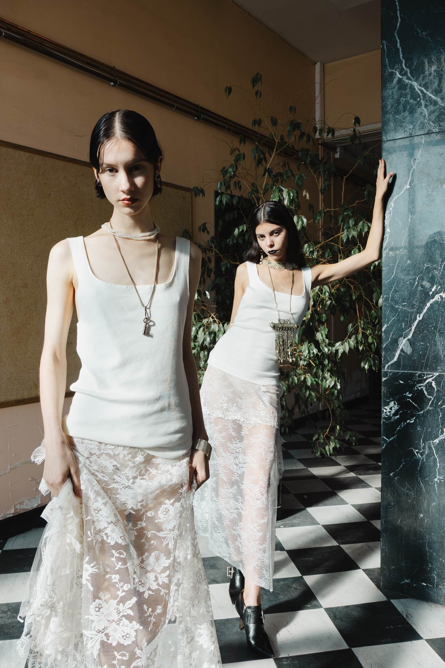 Deux femmes portant des robes en dentelle blanche avec des bijoux, dans un intérieur avec un sol en carrelage noir et blanc et un mur en marbre vert, posant pour une séance photo.