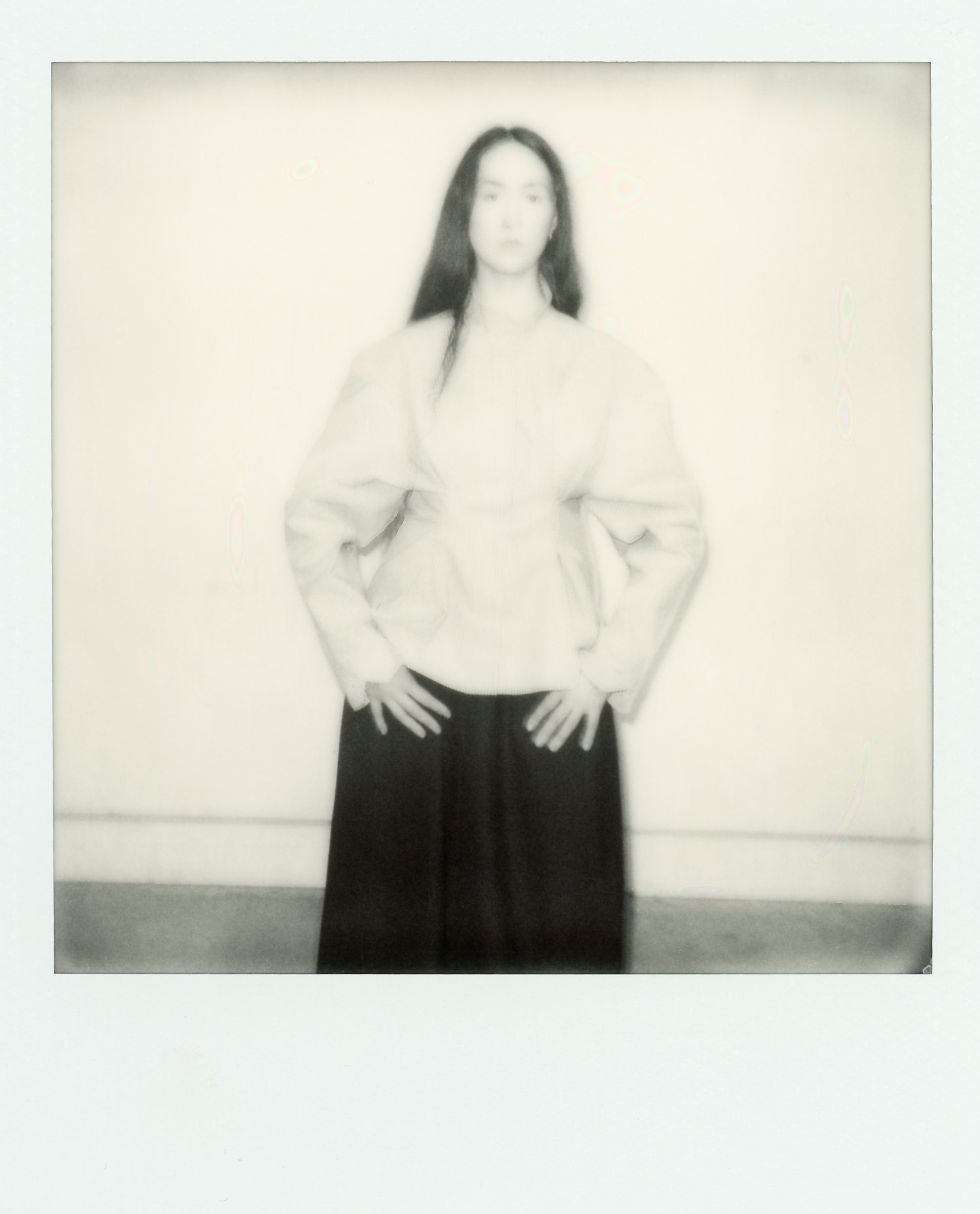 Femme debout, mains sur les hanches, habillée d'une veste claire et d'une robe noire longue, posant devant un mur blanc.