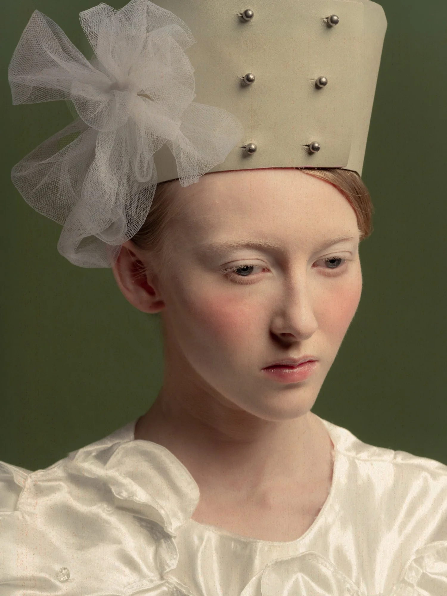 Jeune femme avec un chapeau beige décoré d'un grand nœud en tulle blanc, portant une robe en satin beige, sur fond vert