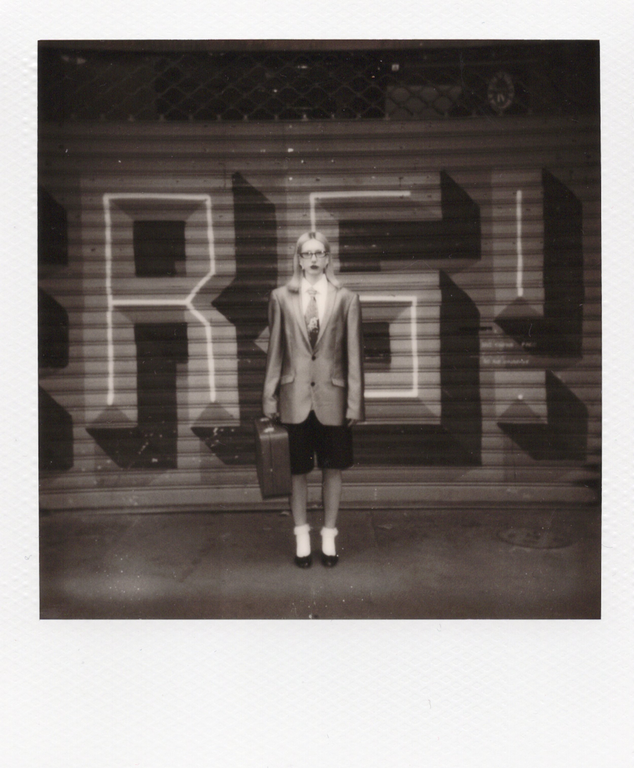 Une personne debout devant un panneau avec des lettres formant le mot 'Berlin', portant une veste longue, une jupe ou short, des chaussettes hautes blanches et des talons, tenant une valise.