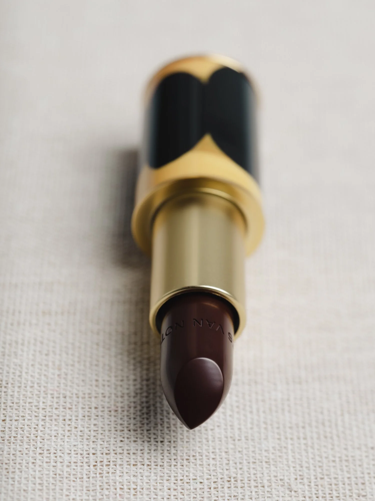 Rouge à lèvres marron foncé avec emballage beige et noir, posé sur un fond blanc.
