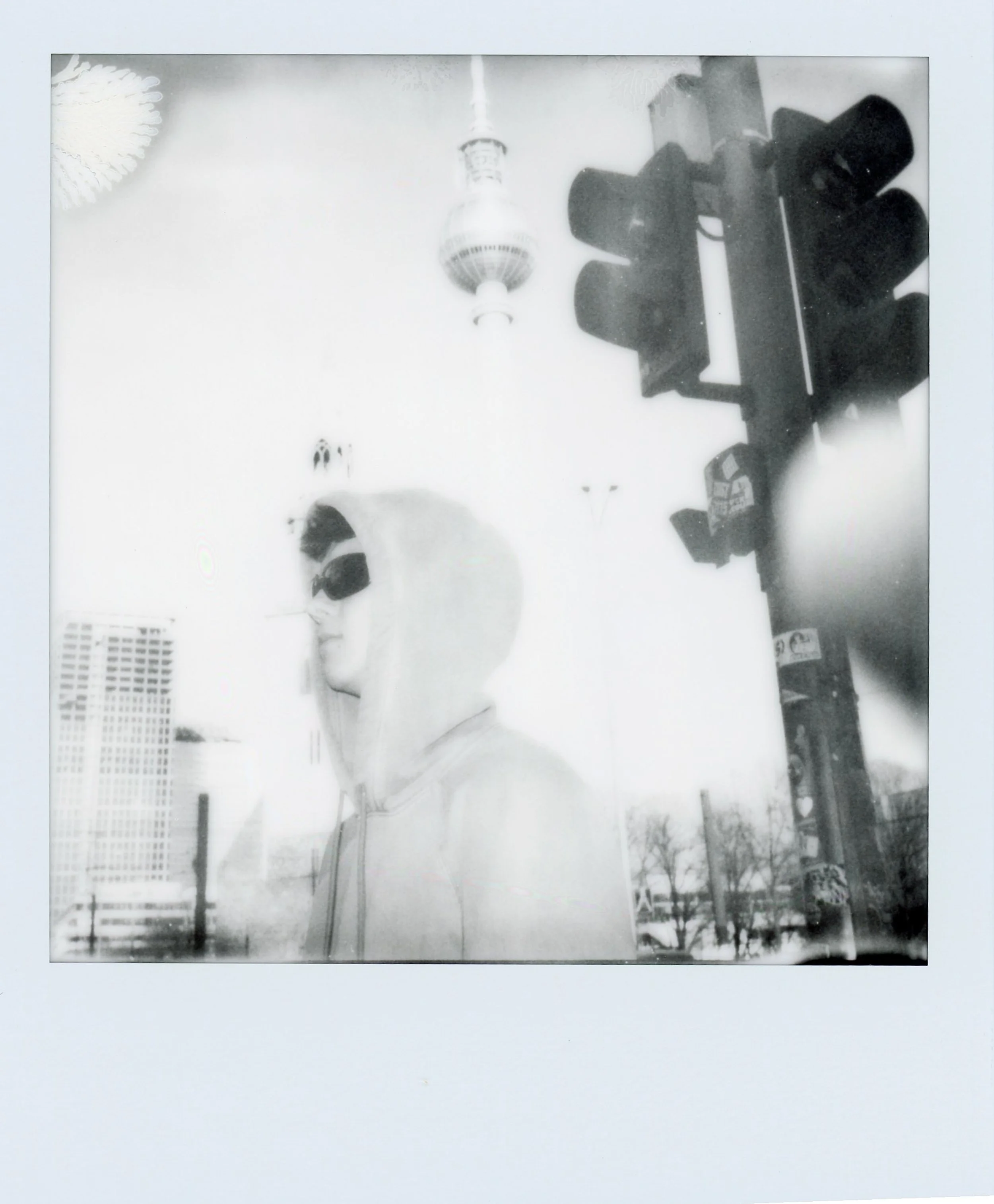 Une personne portant une capuche et des lunettes de soleil, vue à travers une vitre, avec un feu de signalisation à côté et une tour en arrière-plan.