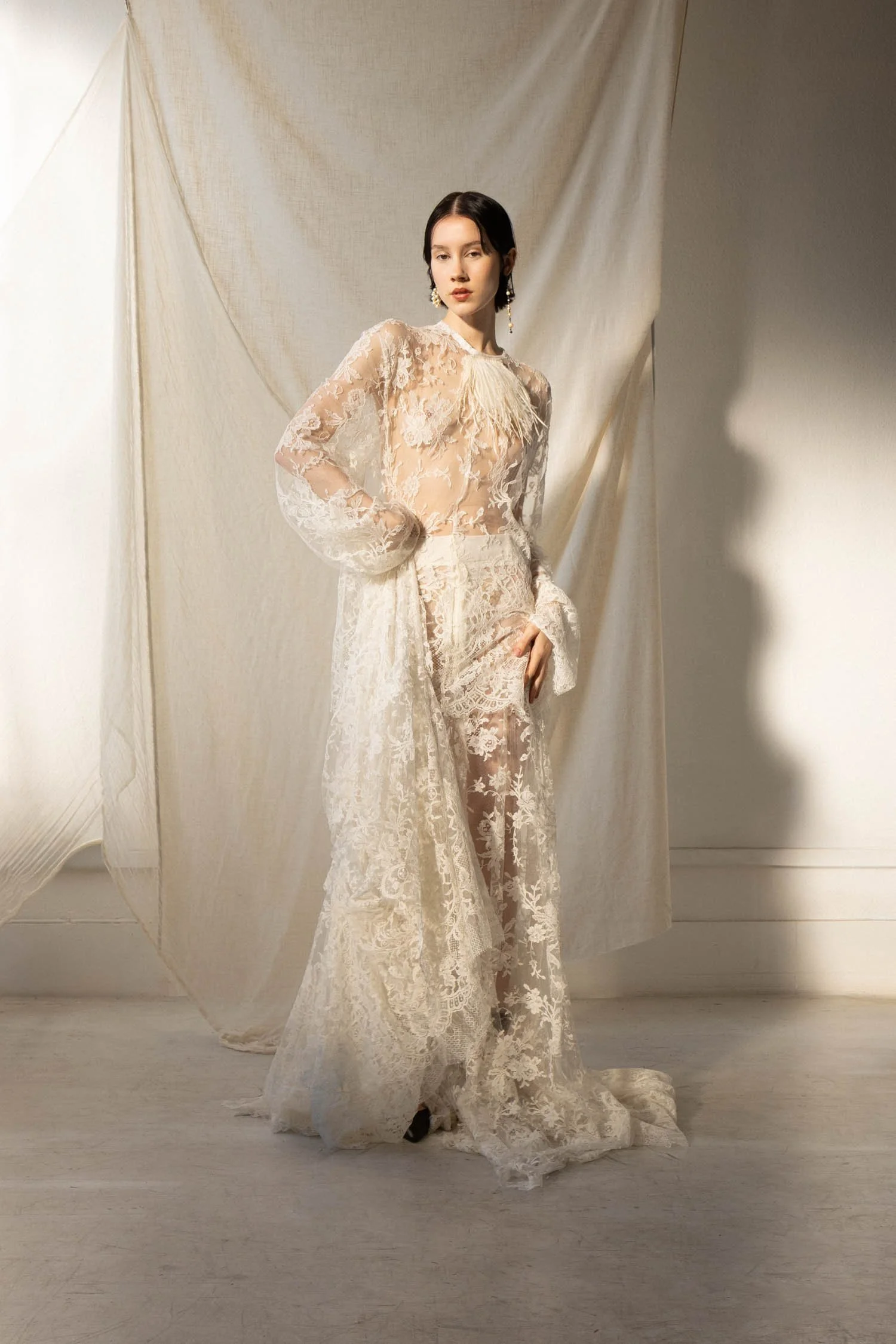 Une femme porte une robe de mariée en dentelle beige clair avec des détails floraux, devant un fond en tissu ivoire.