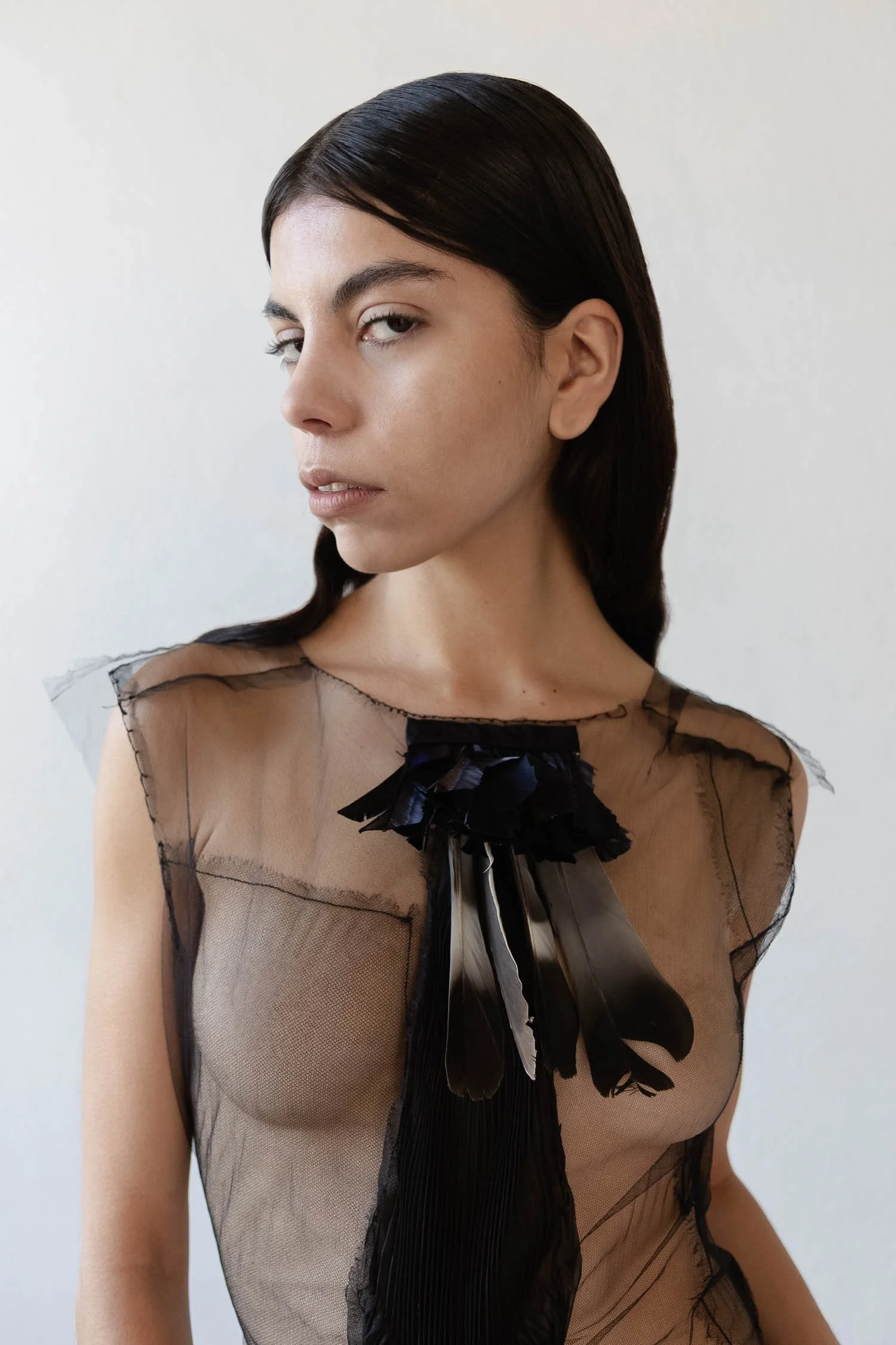 Jeune femme avec des cheveux bruns, vêtue d'une robe en tulle noir transparente avec un détail en plumes noires au col.