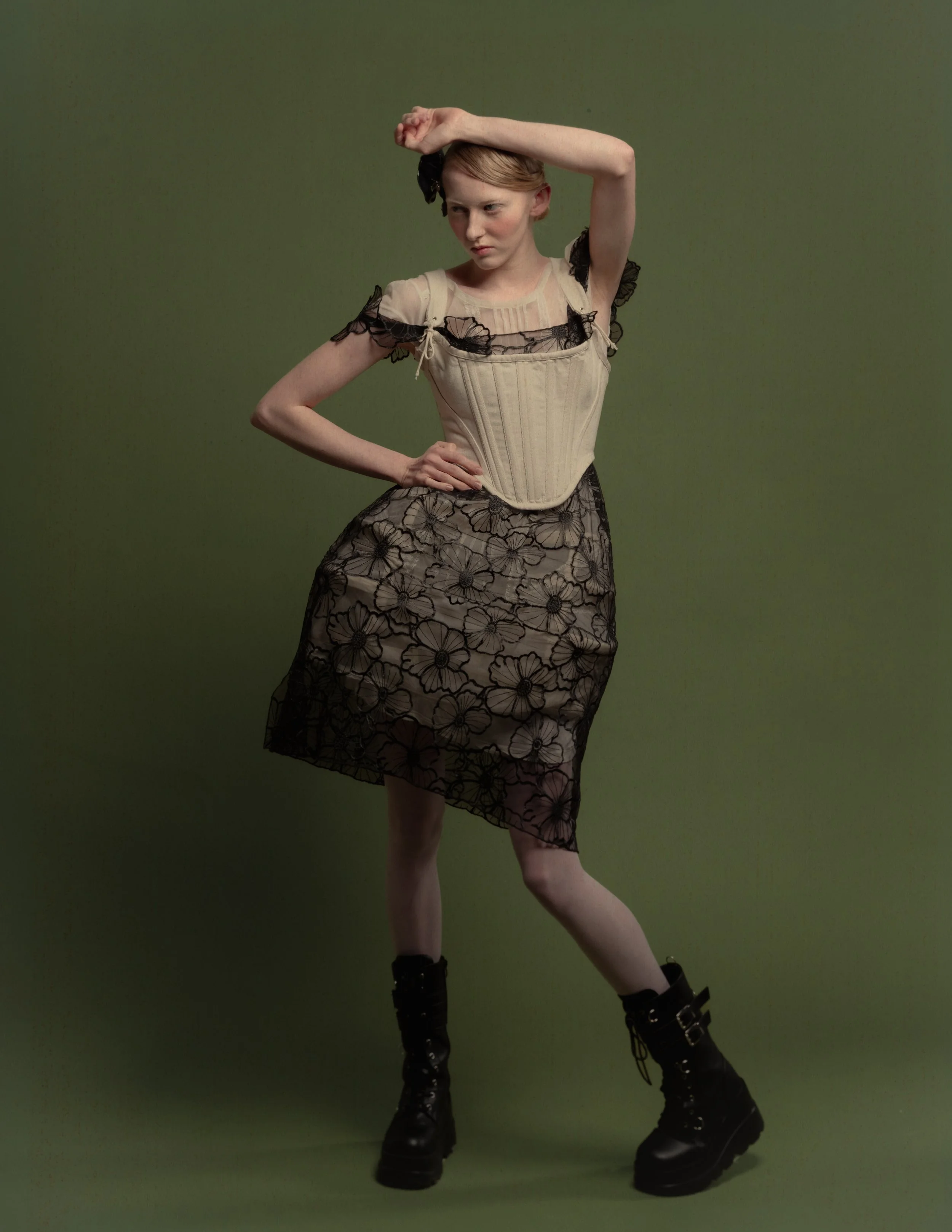 Une femme posant contre un fond vert, portant une robe noire en dentelle, des bottes noires, et un haut beige corseté.