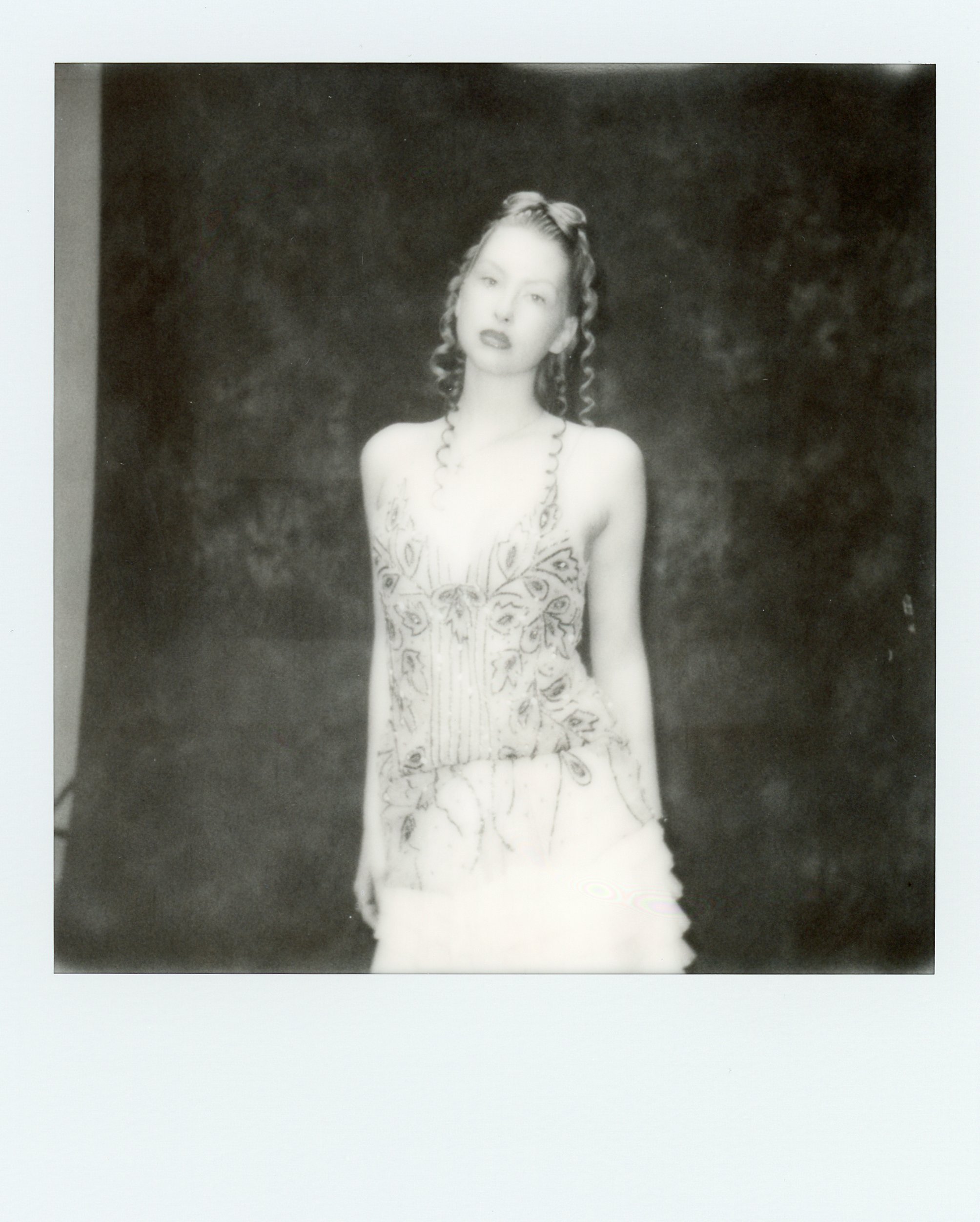 Une jeune femme posant devant un fond sombre, portant une robe légère à motifs et des boucles d'oreilles longues, avec les cheveux bouclés.