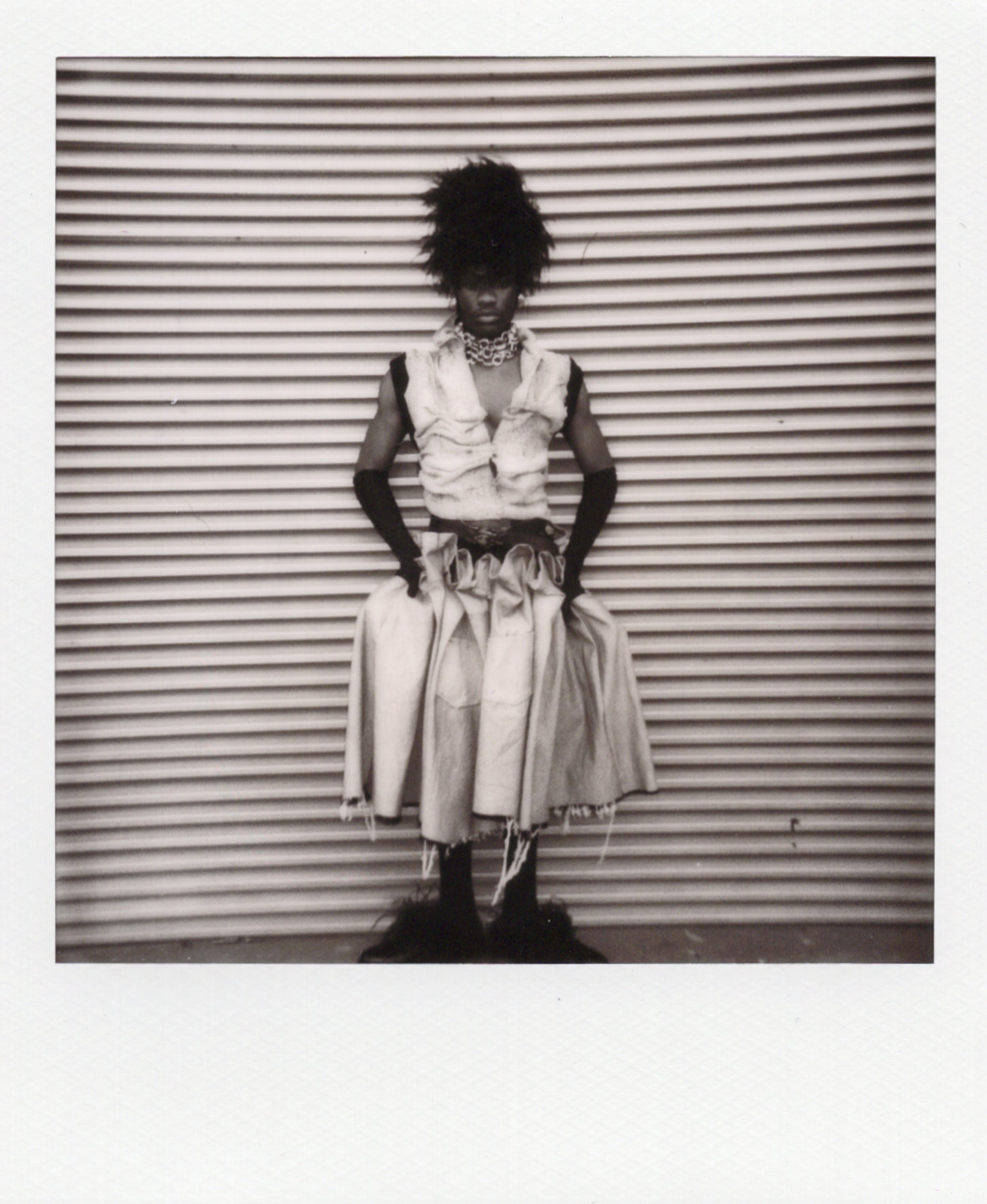 Une femme portant une tenue de mode artistique, avec une jupe ample et un haut sans manches, se tenant devant un mur en métal, avec des accessoires de bijoux et des gants longs noirs.