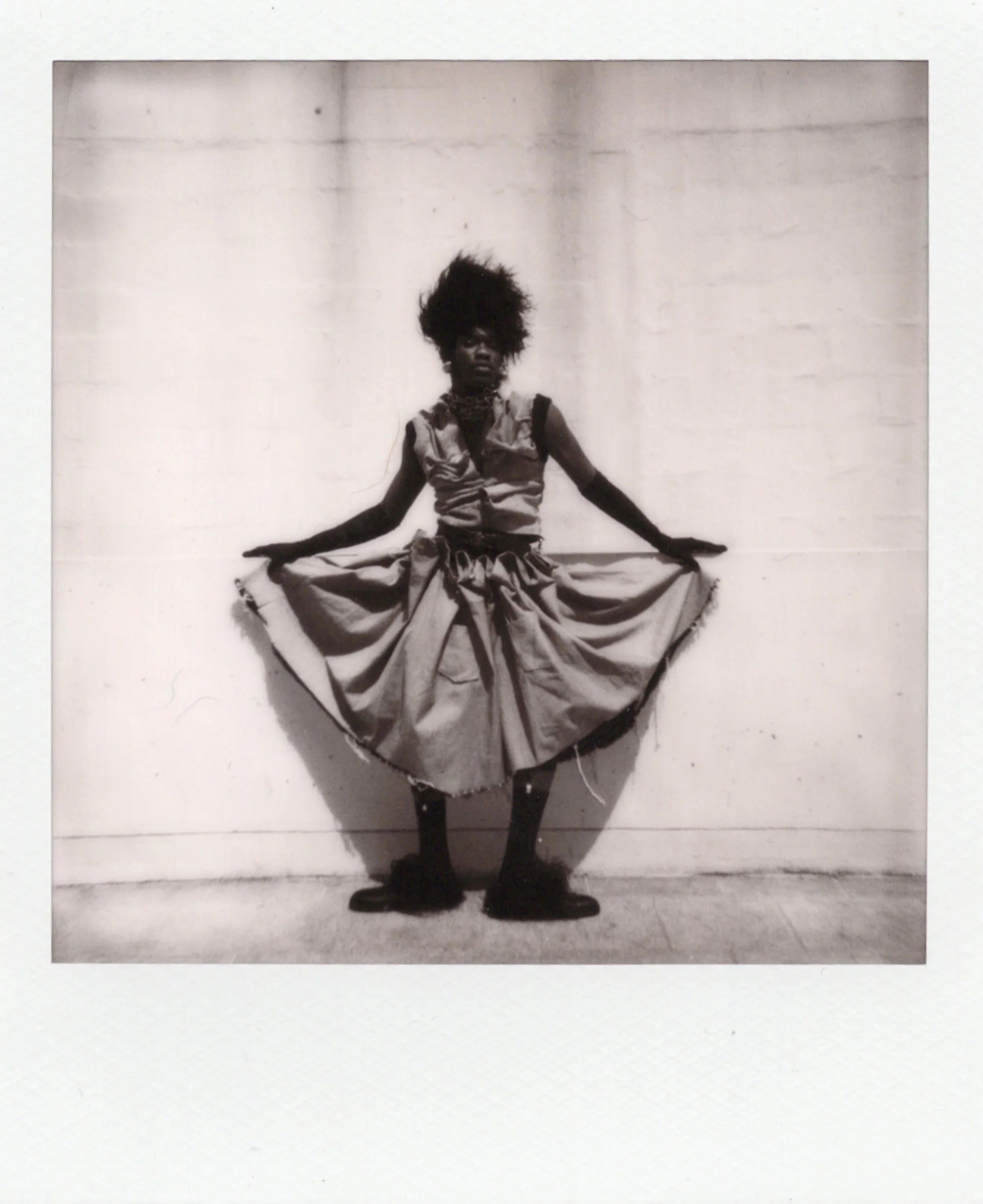 Une femme pose contre un mur, portant une jupe ample et une veste sans manches, avec une coiffure volumineuse et une expression sérieuse.