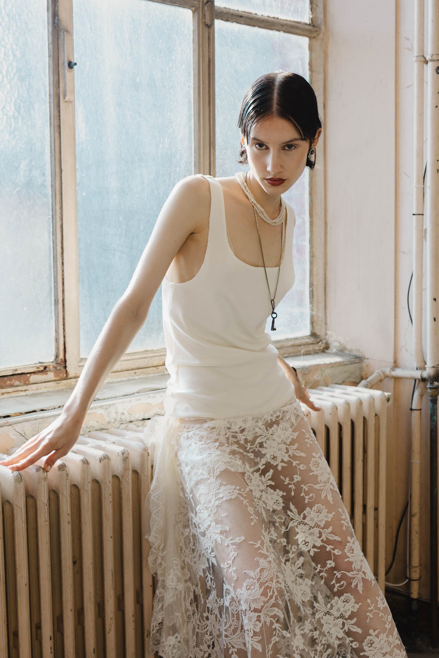 Une jeune femme à la peau claire, aux cheveux noirs courts, porte un haut blanc sans manches et une jupe en dentelle transparente avec un motif floral, posant près d'une fenêtre dans une pièce lumineuse.