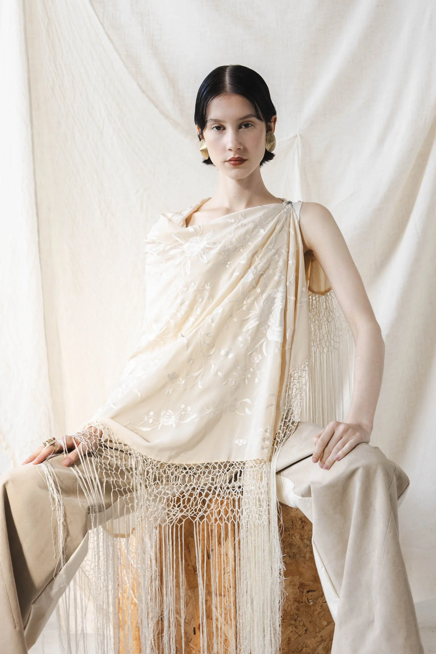 Une femme posant en portant une robe élégante beige avec des broderies blanches et un détail en macramé, avec un fond en tissu beige clair.