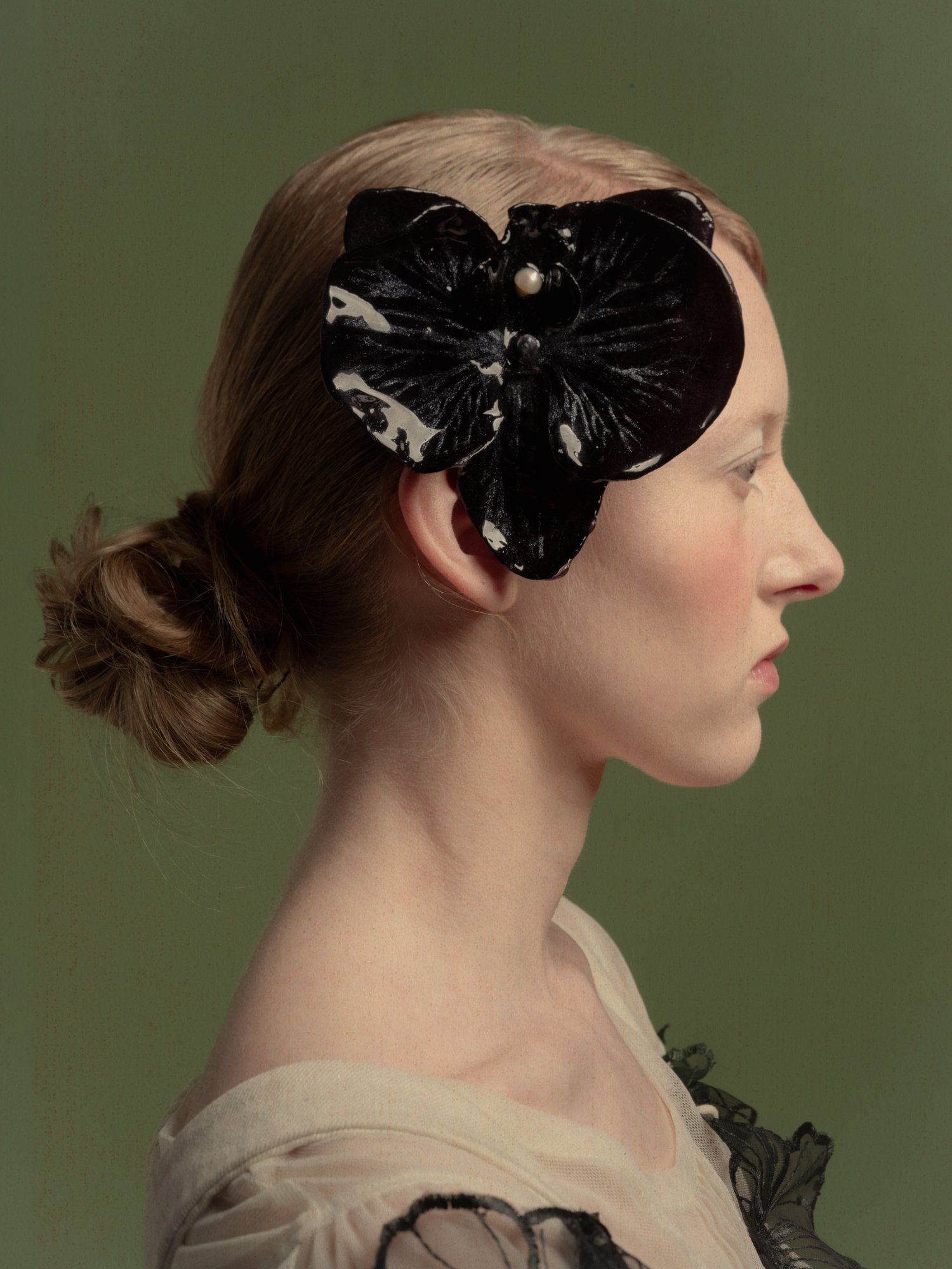 Profil d'une femme aux cheveux blond clair coiffés en chignon, portant un grand accessoire en forme de papillon noir brillant avec des perles, contre un fond vert