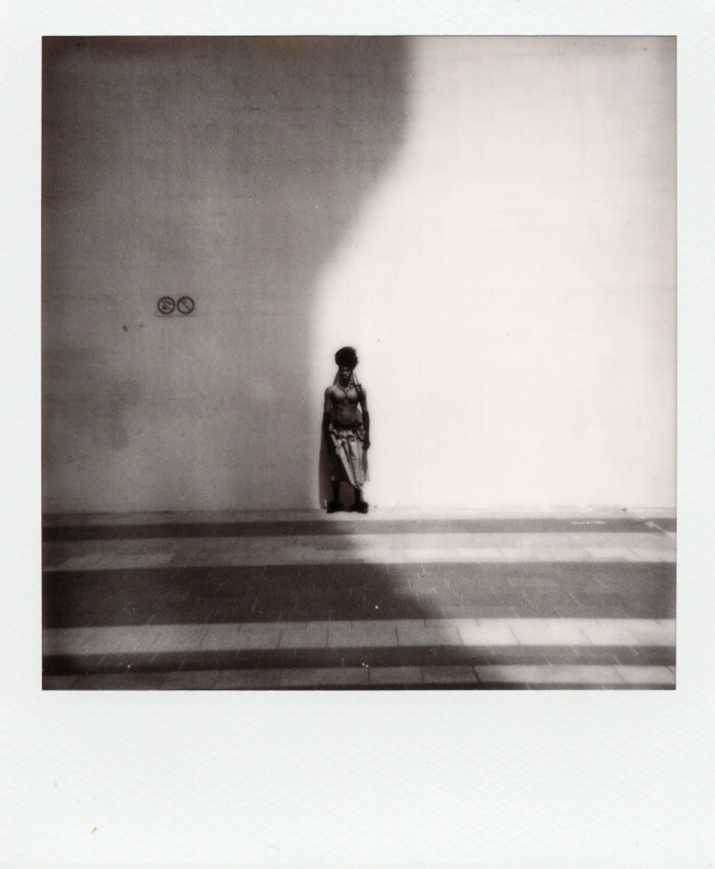 Une femme se tient devant un mur blanc, partiellement ombragé, avec des lignes horizontales au sol, dans une photographie en noir et blanc.