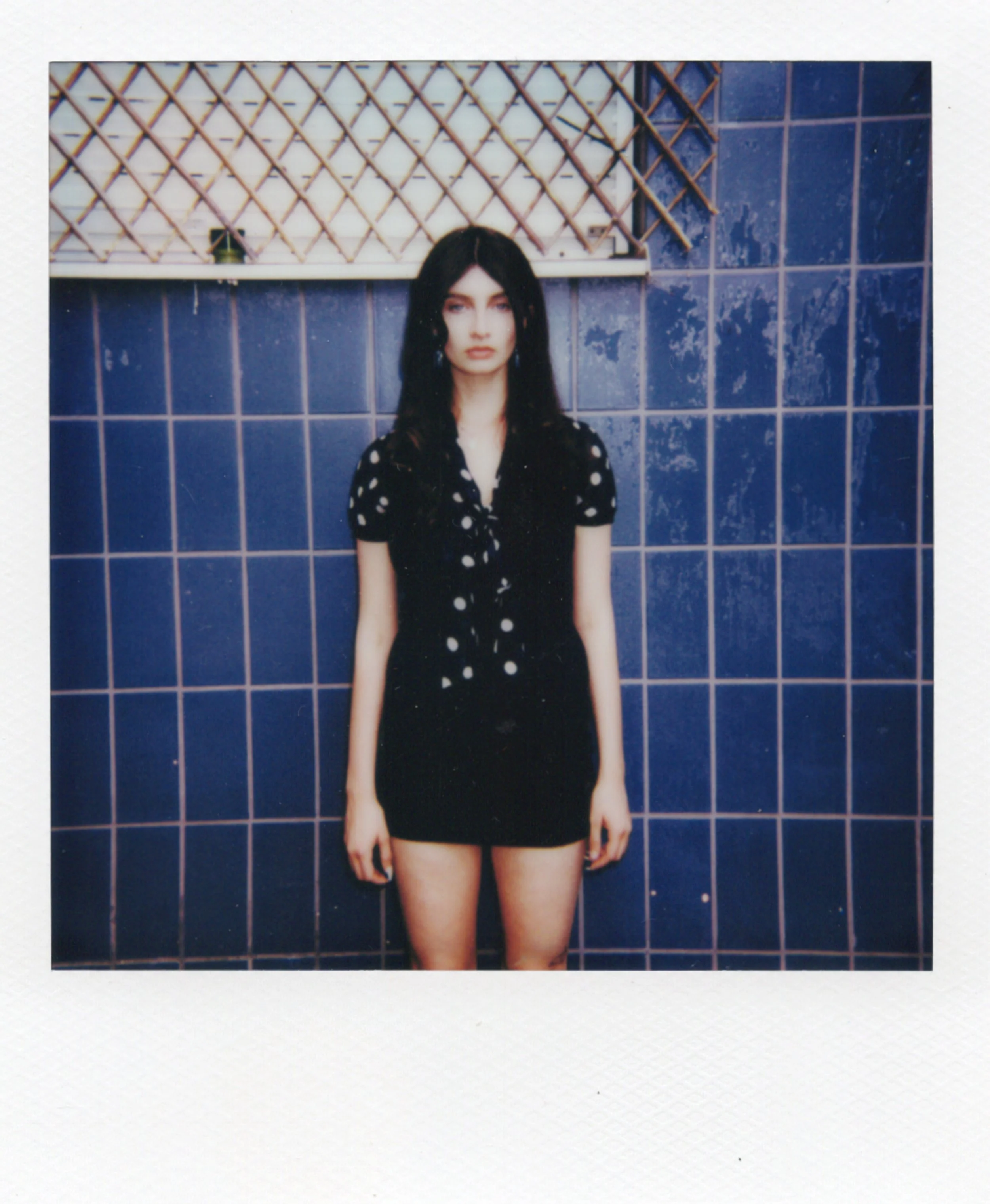 Jeune femme avec des cheveux noirs, portant une robe noire à pois blancs, debout devant un mur en carreaux bleus.