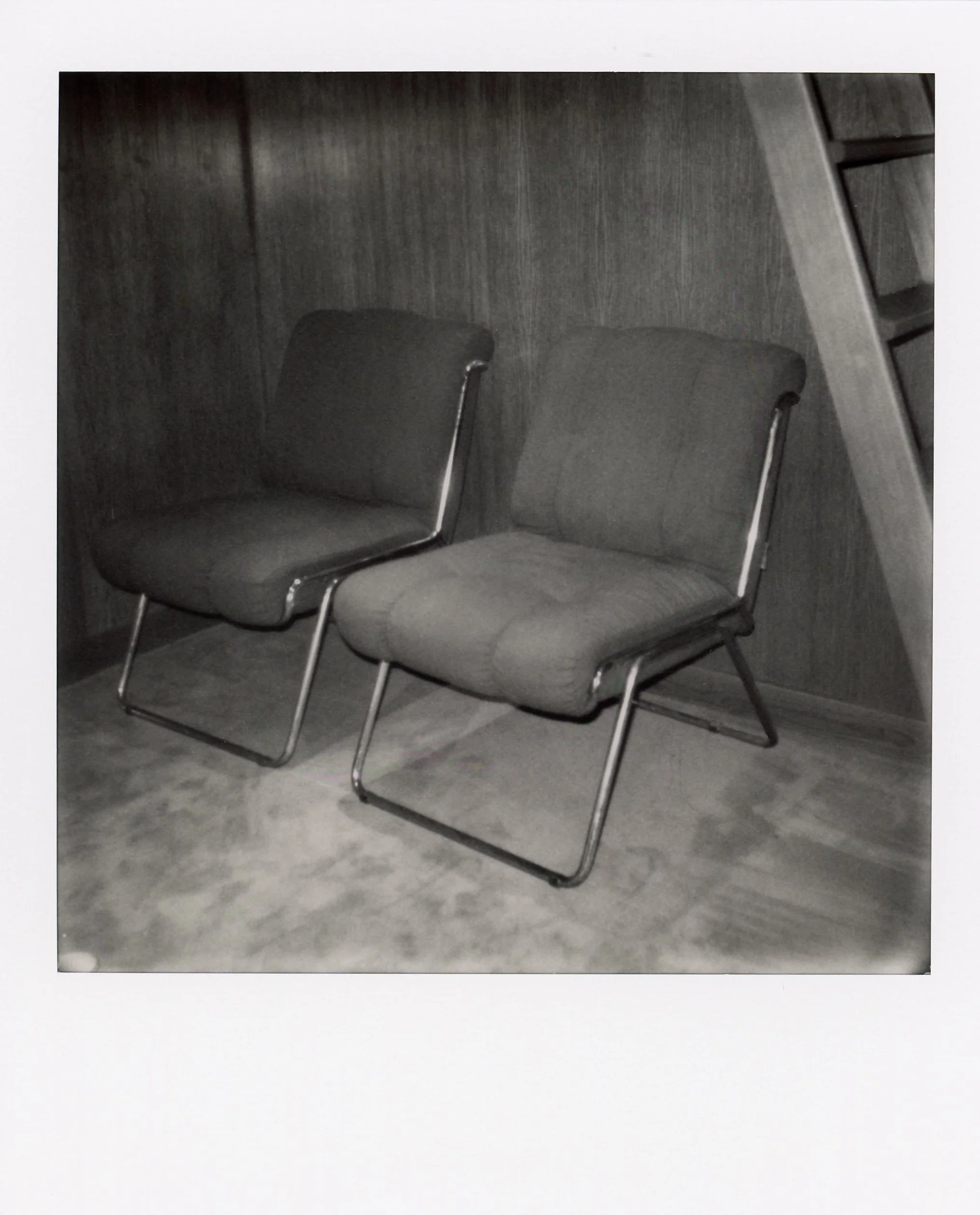 Deux fauteuils en tissu gris avec un cadre métallique, placés sur un tapis dans une pièce au mur en bois foncé, avec un escalier en bois adjacent.