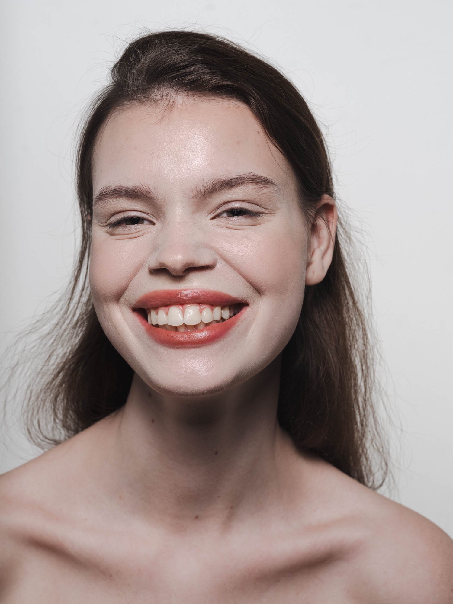 Un portrait d'une femme souriante avec les épaules dénudées, cheveux bruns, et teint clair, contre un fond blanc.