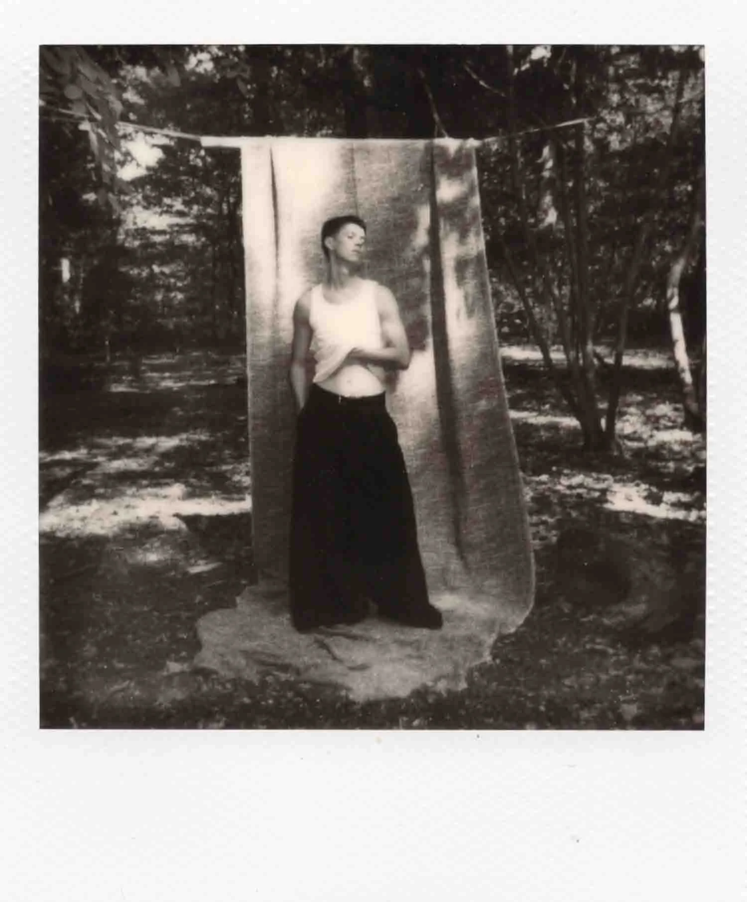 Une jeune femme pose devant un rideau suspendu dans un environnement extérieur boisé, en noir et blanc.