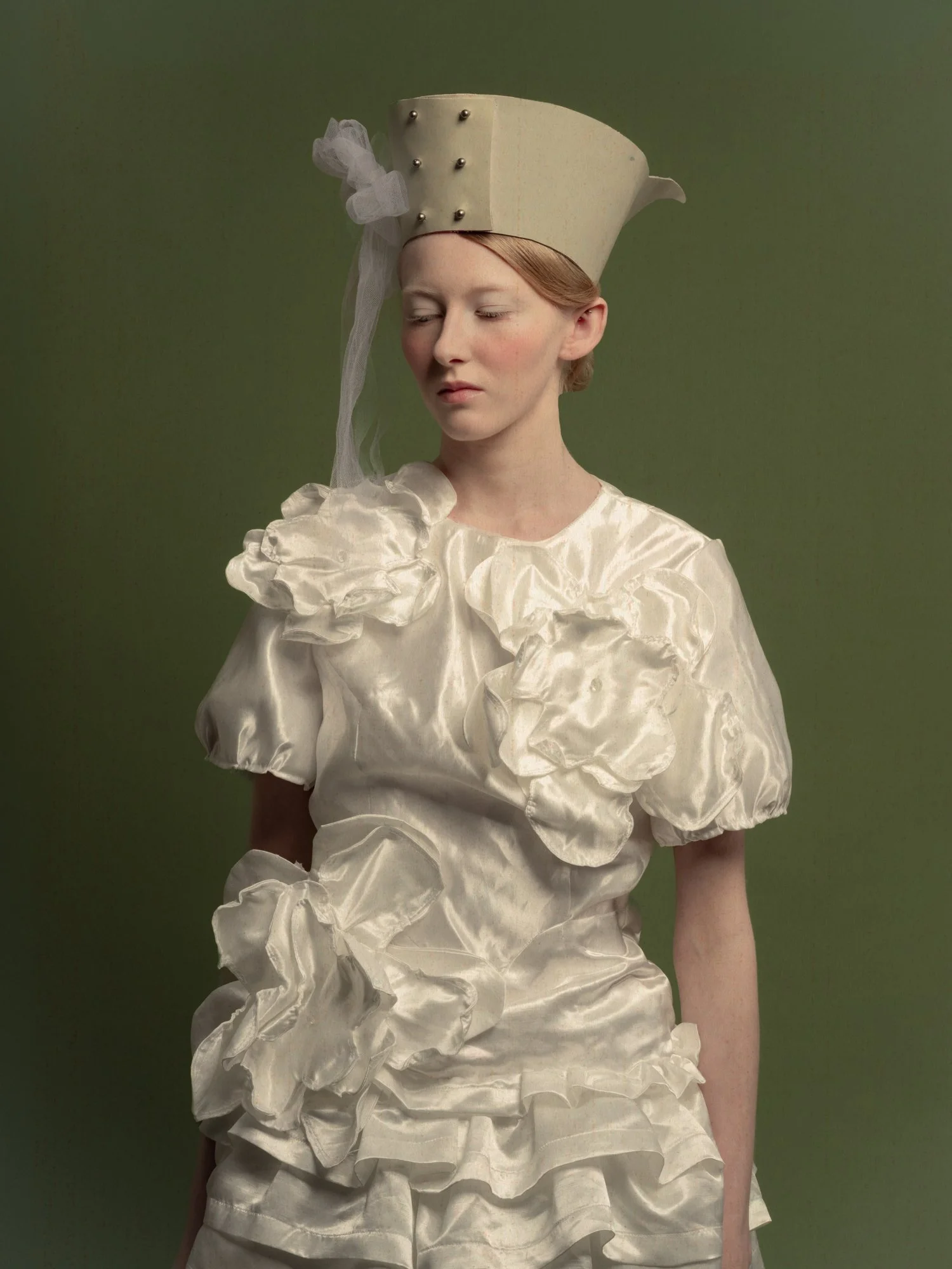 Une femme portant une robe blanc satinée avec des détails en forme de fleurs et un chapeau blanc orné de perles, avec un fond vert.