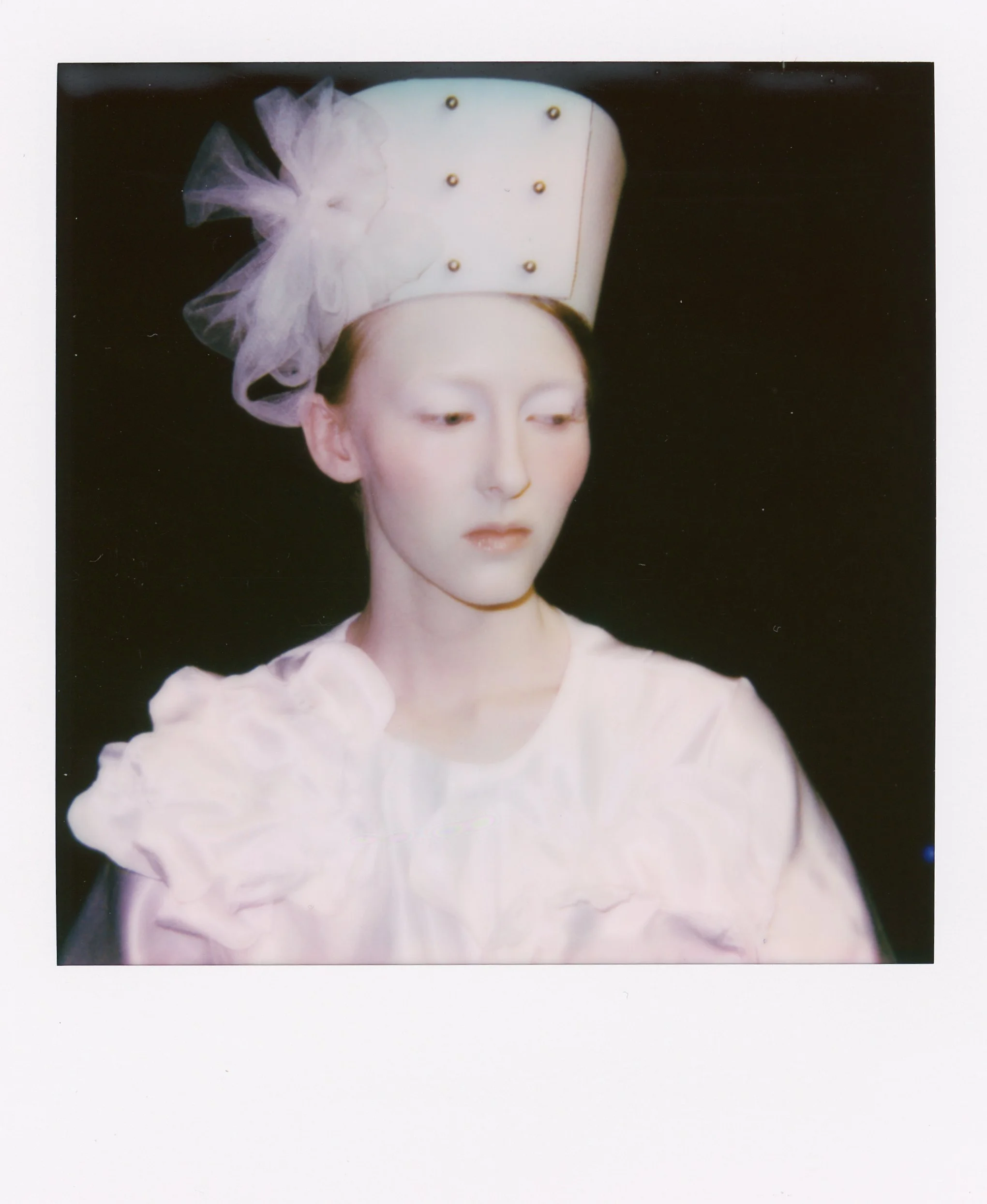 Portrait d'une femme portant un chapeau blanc orné d'une décoration en tulle et de perles, vêtue d'une robe blanche.