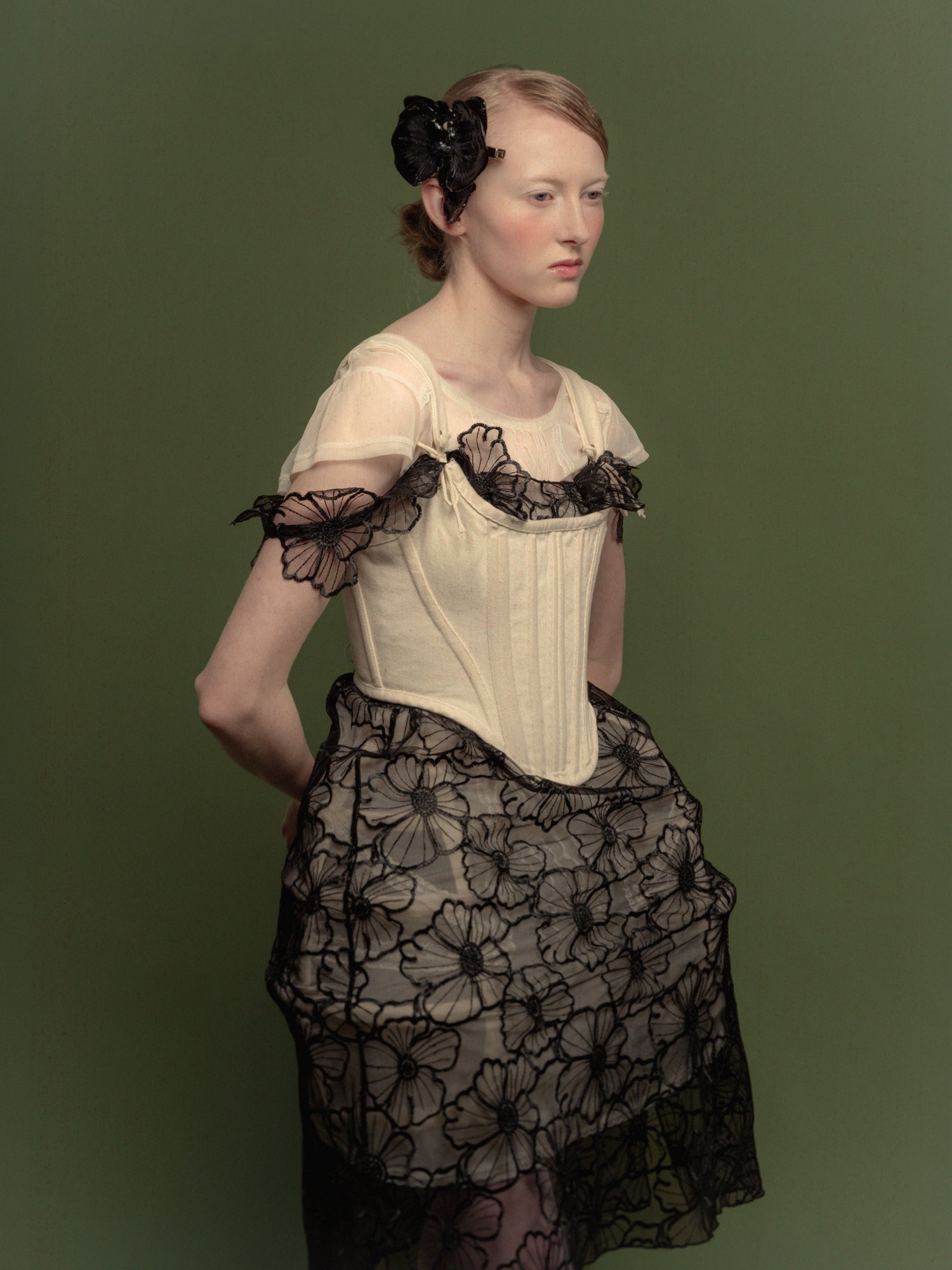 Une femme portant une robe verte avec un corsage beige et des détails en dentelle noire, côtoyée d'une grande fleur noire dans ses cheveux.