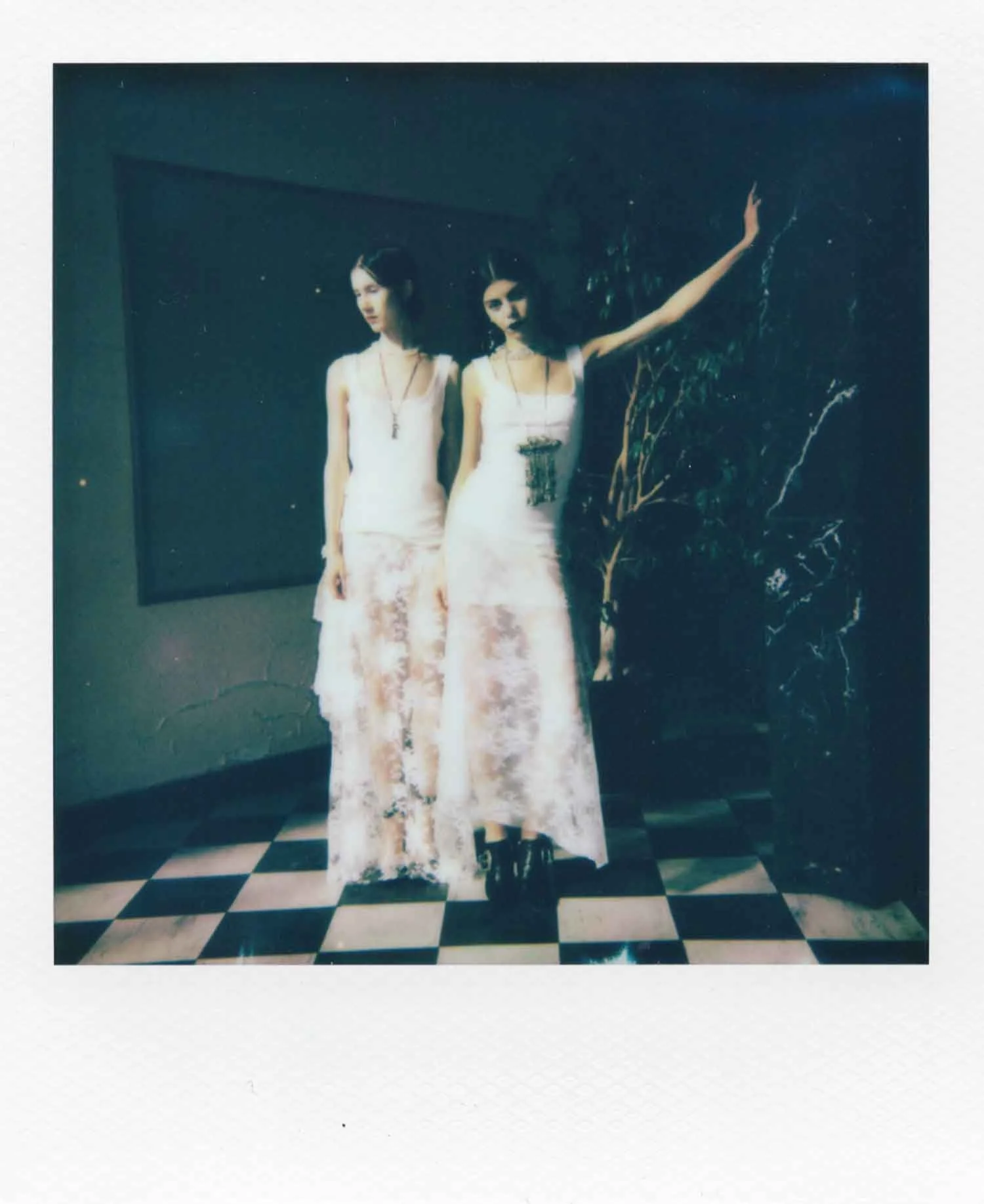 Deux jeunes femmes en robes longues debout sur un sol en carreaux de parole, près d'un végétal, dans une pièce sombre, avec un tableau noir en arrière-plan.