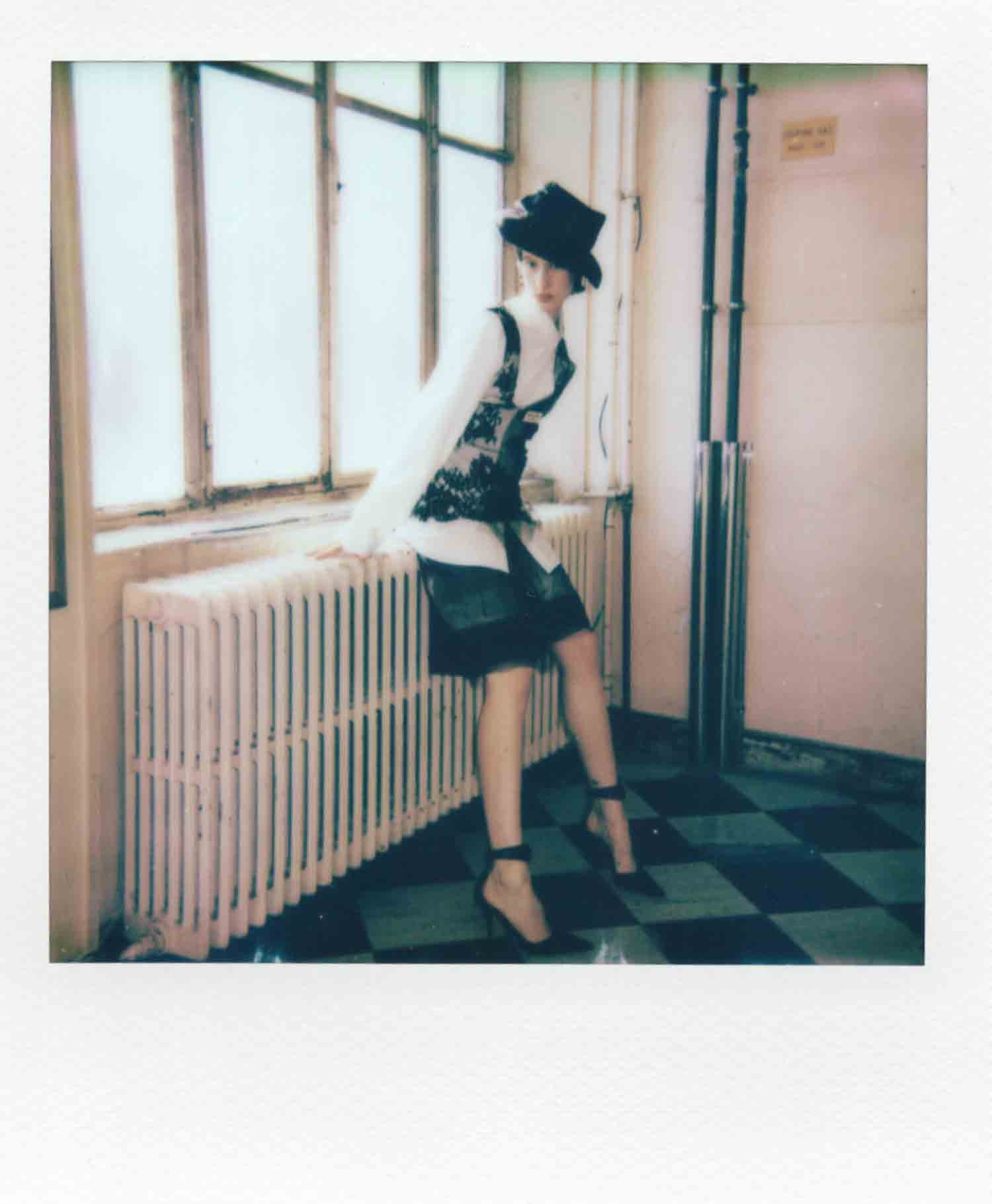 Une femme portant un chapeau noir, un pull blanc et une veste avec un motif noir, assise sur un radiateur dans une pièce avec fenêtre et sol à carreaux.