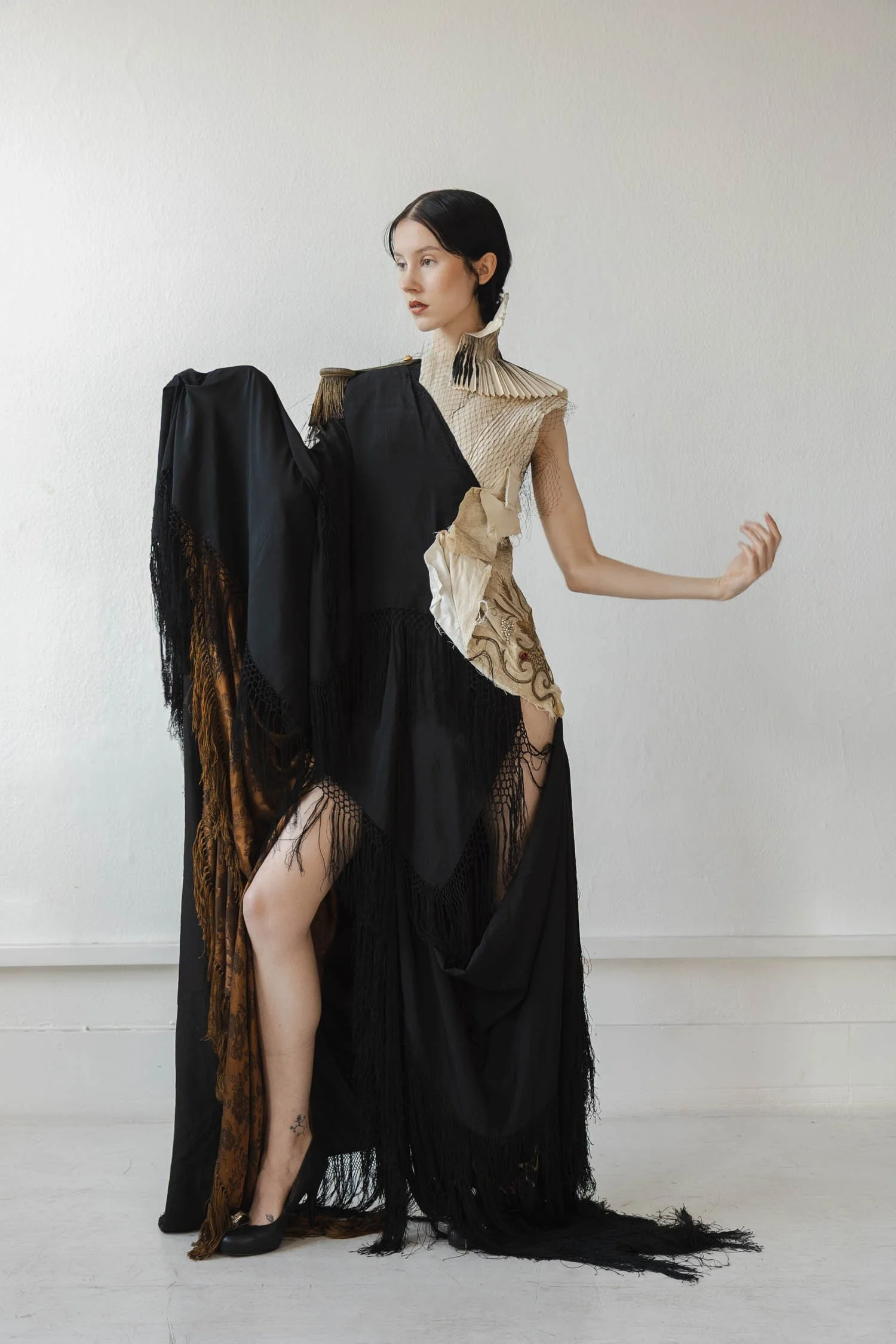 Une femme en robe de créateur mixant tissus noirs et beige, avec des éléments en plumes et broderies, positionnée devant un mur blanc.