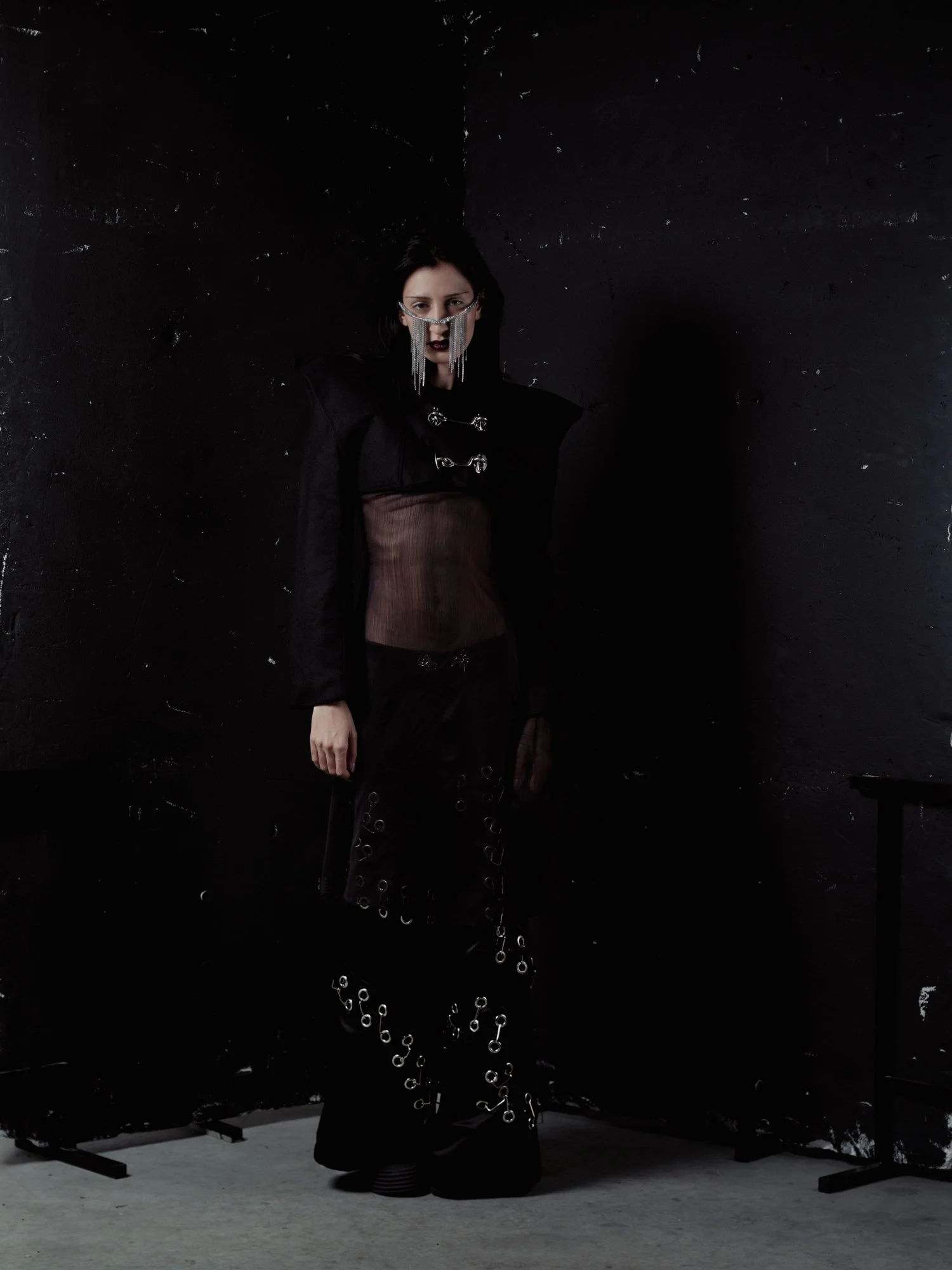 Une femme habillée en tenue noire avec des accessoires métalliques, portant des lunettes avec des chaînes, se tenant devant un mur noir dans une ambiance sombre et gothique.