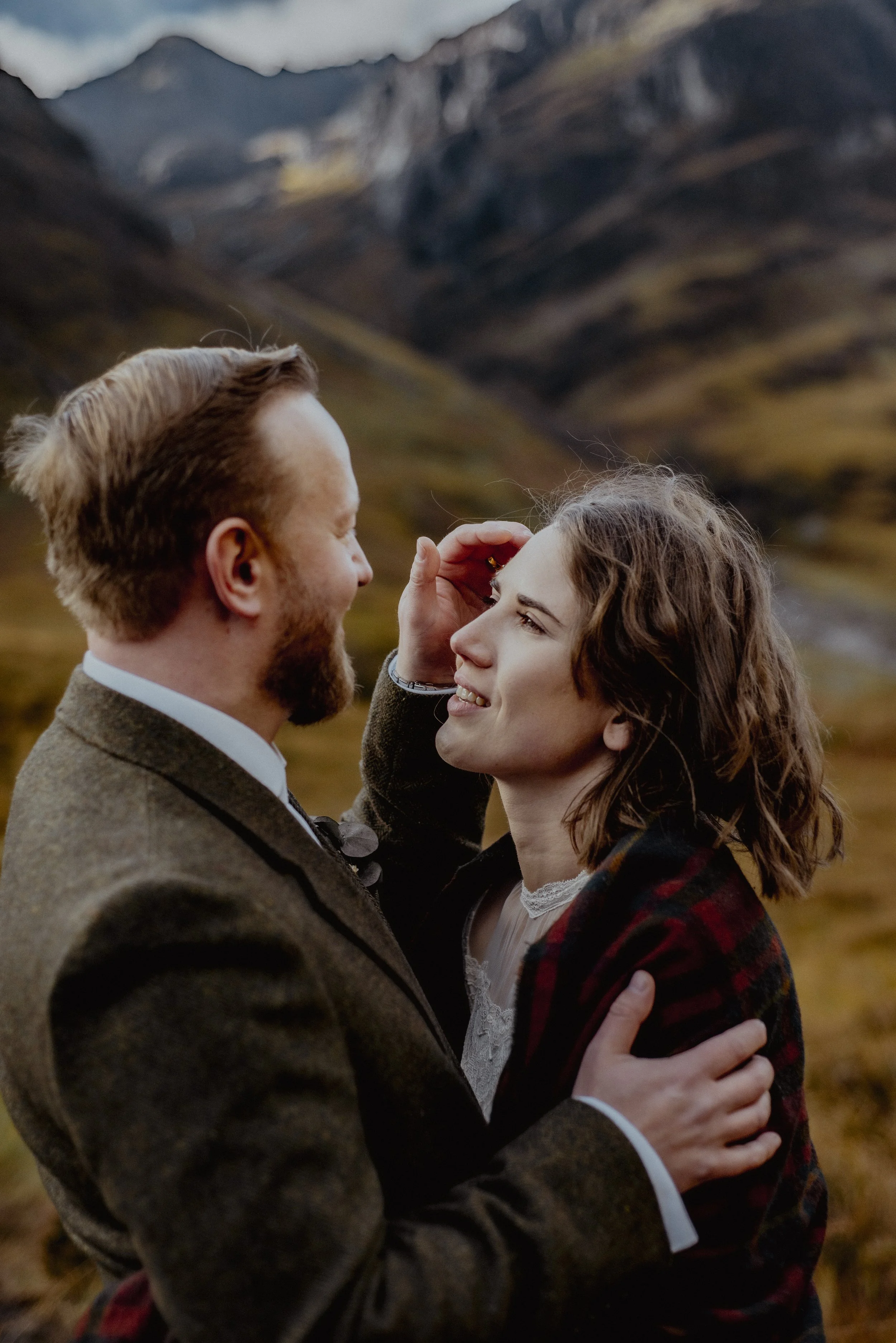 A couple smiling and embracing outdoors in a mountainous landscape, with the man touching the woman's forehead.
