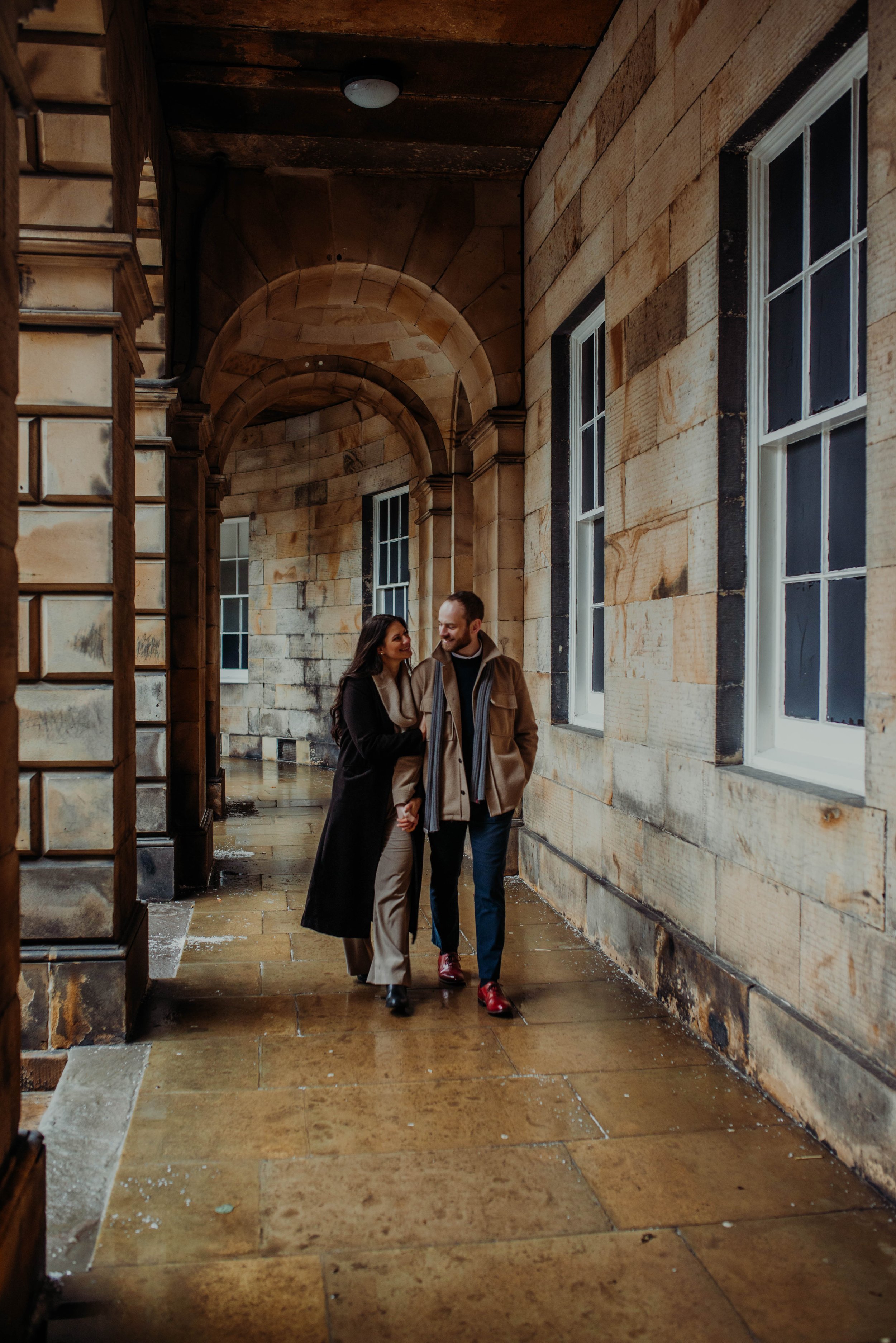 A couple walking and talking in a stone hallway with arched ceilings and multiple windows, during a rainy day.