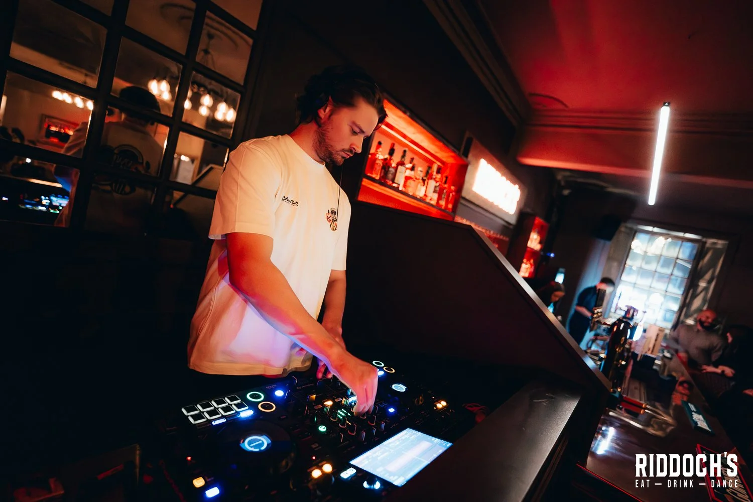 DJ performing at a bar, surrounded by bottles and bar equipment, with patrons in the background, in a dimly lit setting.