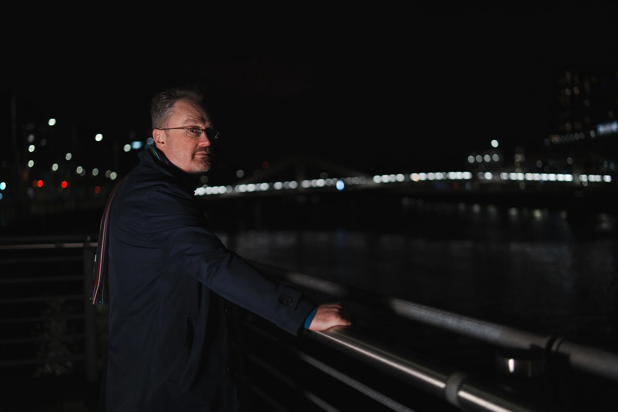 Daniel Sellers with glasses on and a dark coat standing by a railing at night, overlooking a river with a city bridge and illuminated buildings in the background.