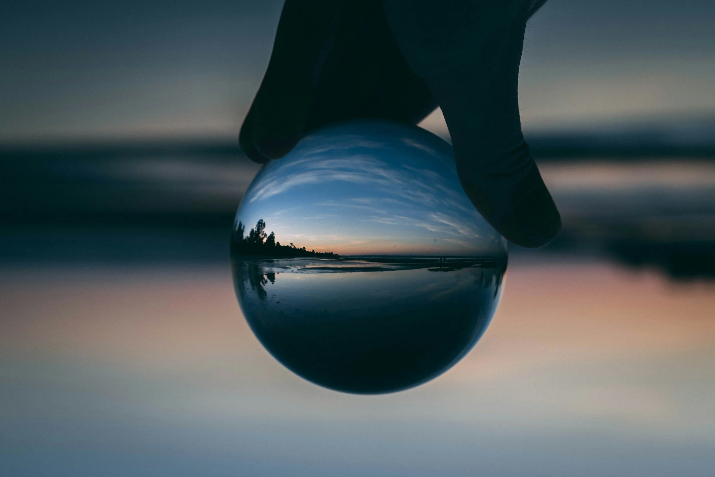 A person holding a glass ball in their hand at the beach during sunset, reflecting the sky and shoreline.